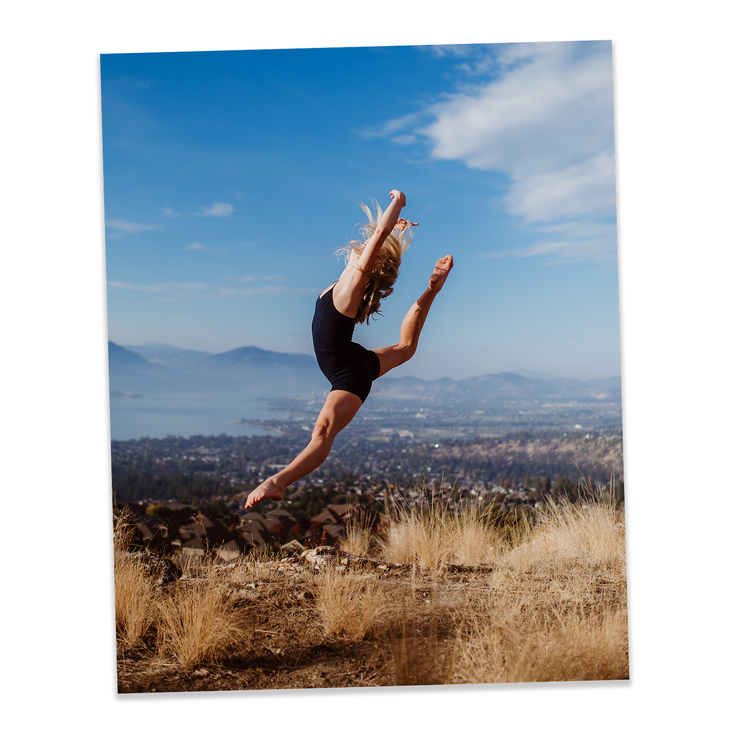 Dancer leaps gracefully through air against mountainous landscape and blue sky backdrop with dry grass in foreground.
