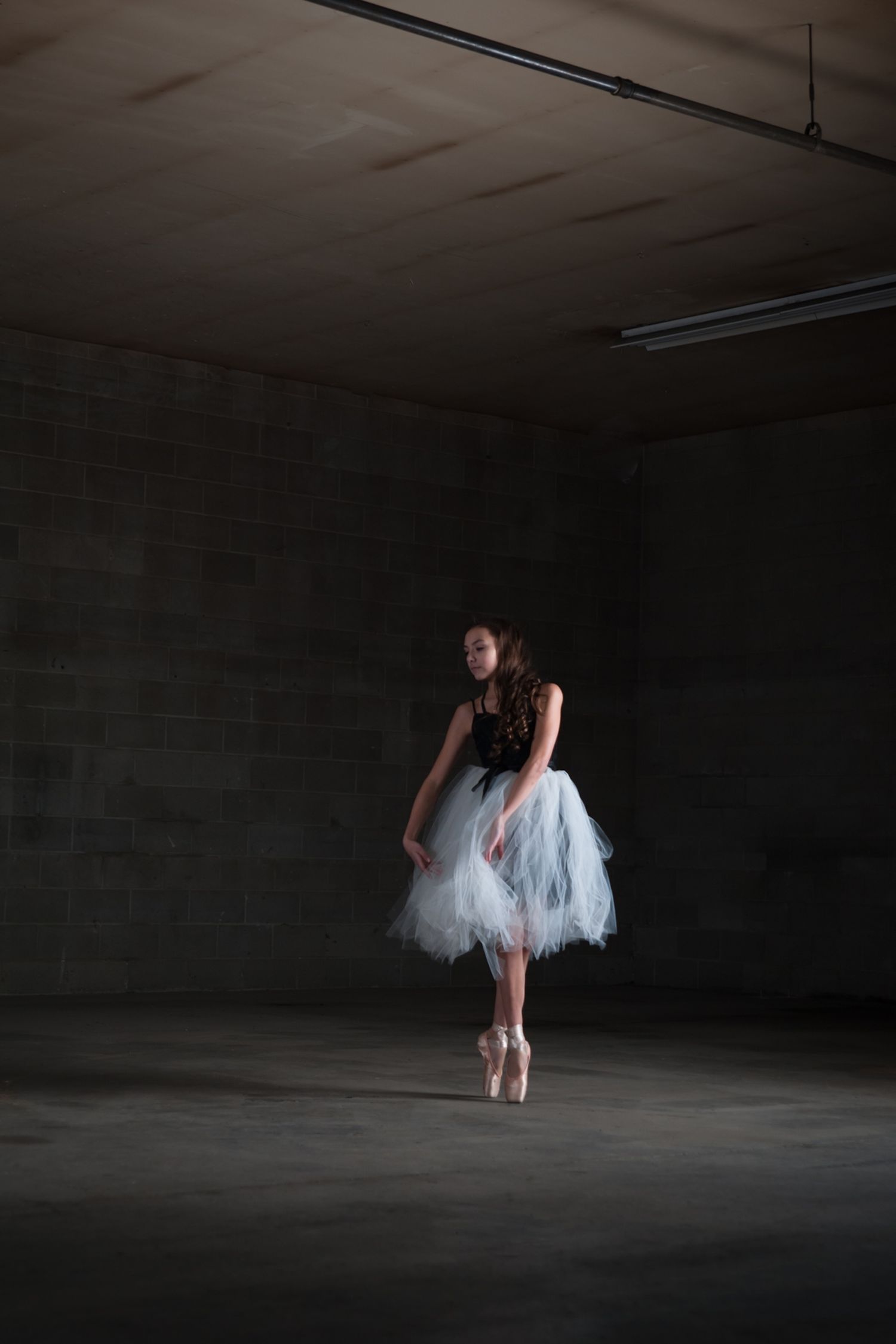A ballet dancer in a white tutu performs en pointe in a dark industrial space.