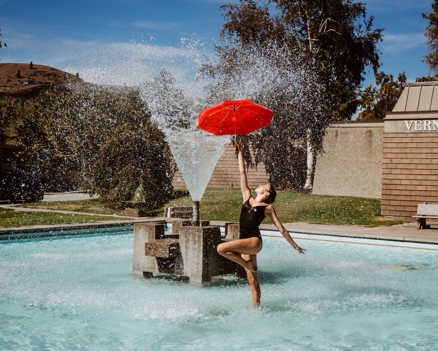 Someone splashes playfully in a fountain holding a bright red umbrella on a sunny day near brick buildings.