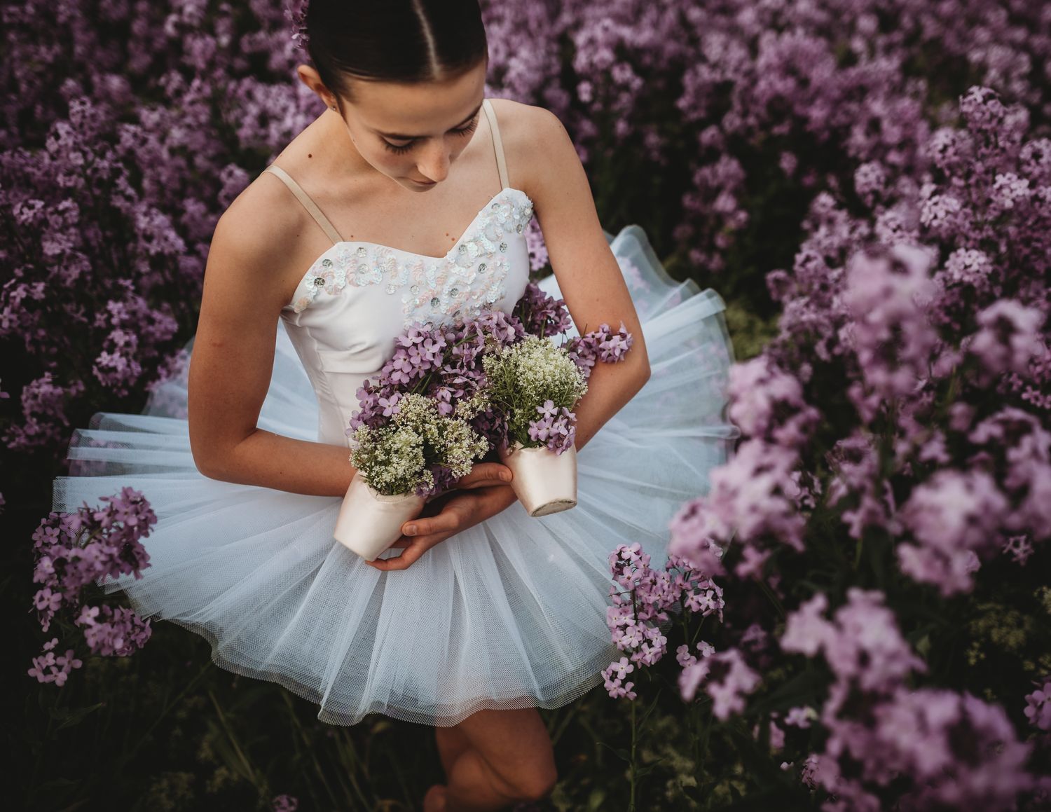 Ballerina in white tutu holding purple wildflowers poses gracefully among blooming lilac bushes in springtime garden.