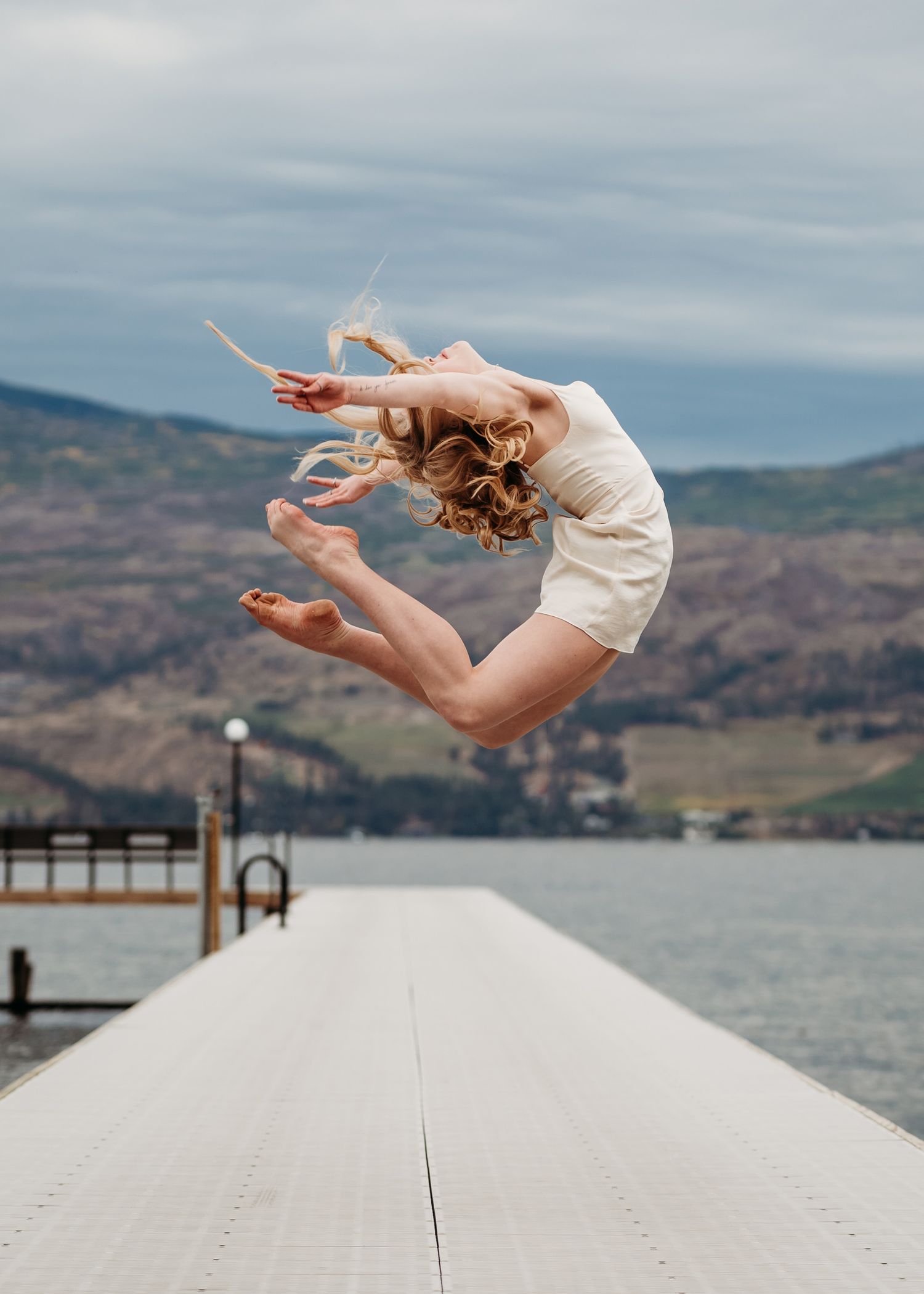 A dancer in white leaps gracefully on a wooden dock over a scenic mountain lake.