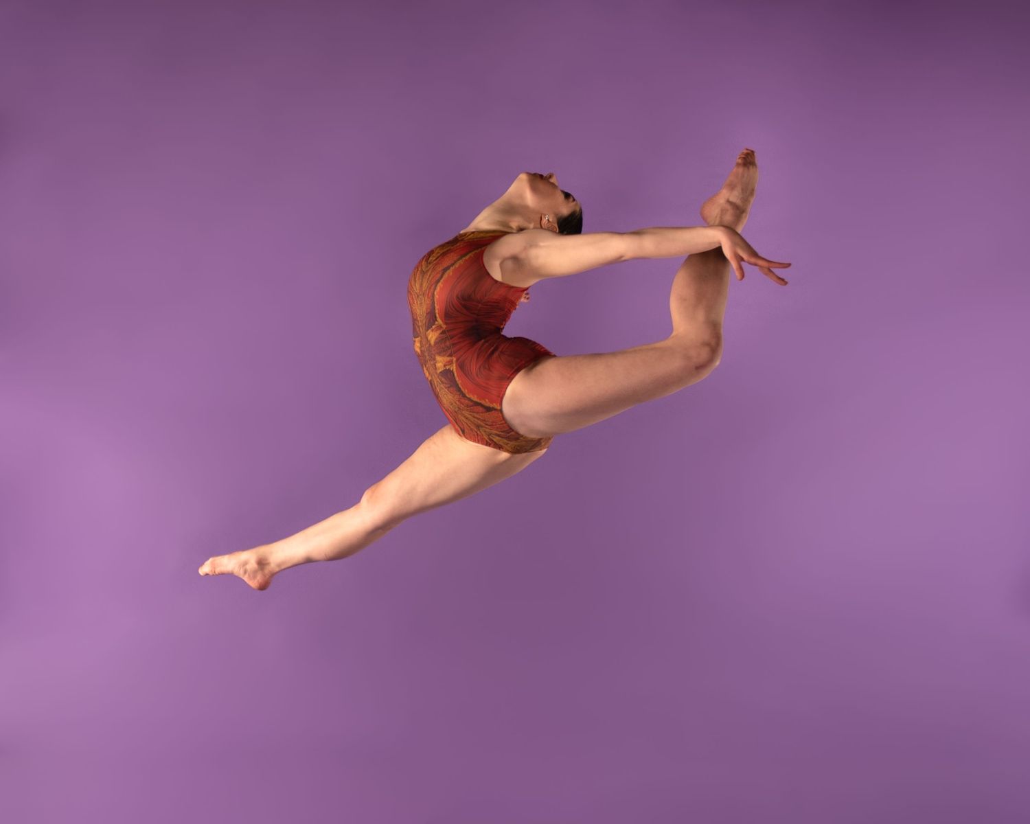 Graceful dancer performs a dramatic split leap against a purple backdrop while wearing a red leotard.