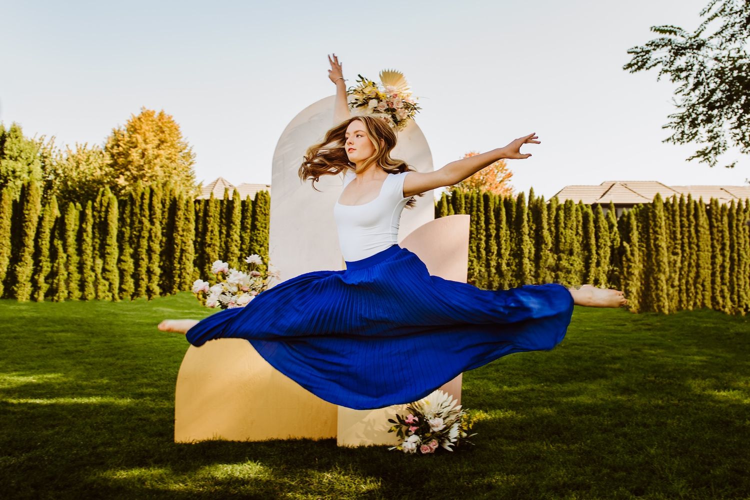 A person in a white top and flowing blue skirt spins joyfully in a sunlit garden with green hedges in the background.