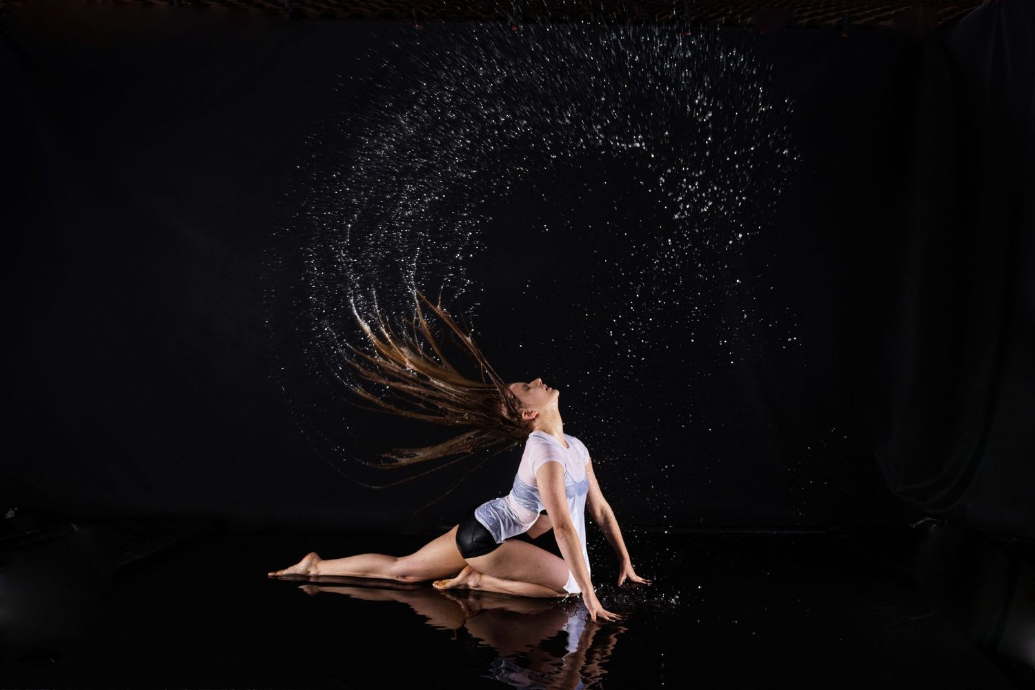 Graceful dancer performs on wet floor with water droplets creating sparkling effect in dramatic lighting.