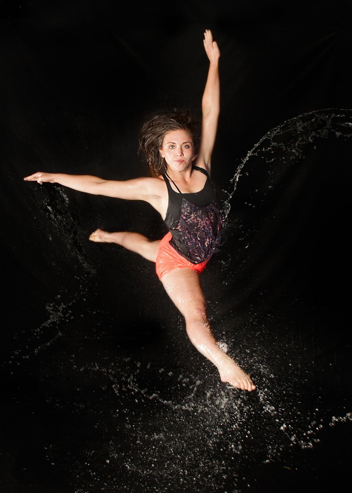 Dynamic dancer in coral shorts and black top leaps through water splashes against dark background creating artistic movement.