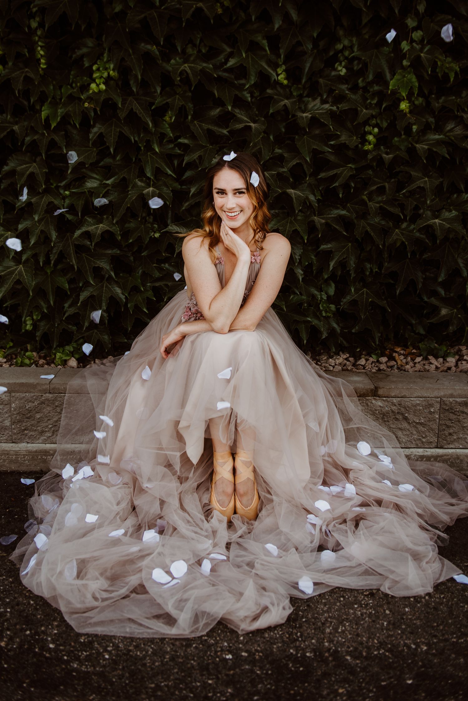 A model in a flowing blush tulle gown sits gracefully against a dark hedge backdrop with falling white flower petals.