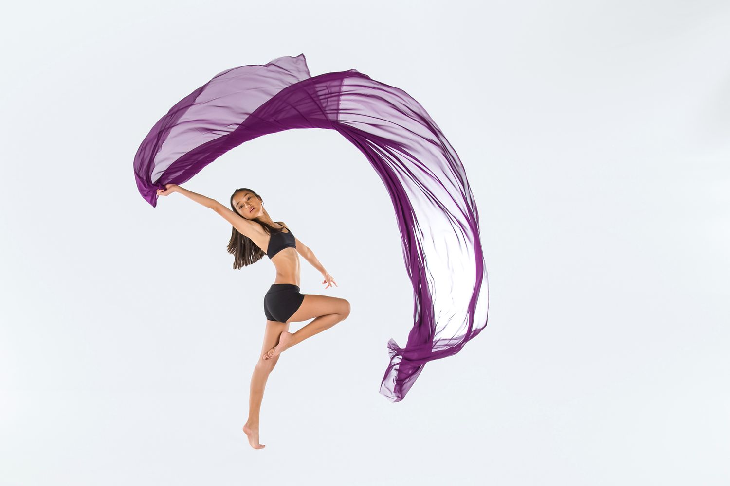 A dancer leaps gracefully through the air with a flowing purple fabric billowing behind them against a white backdrop.