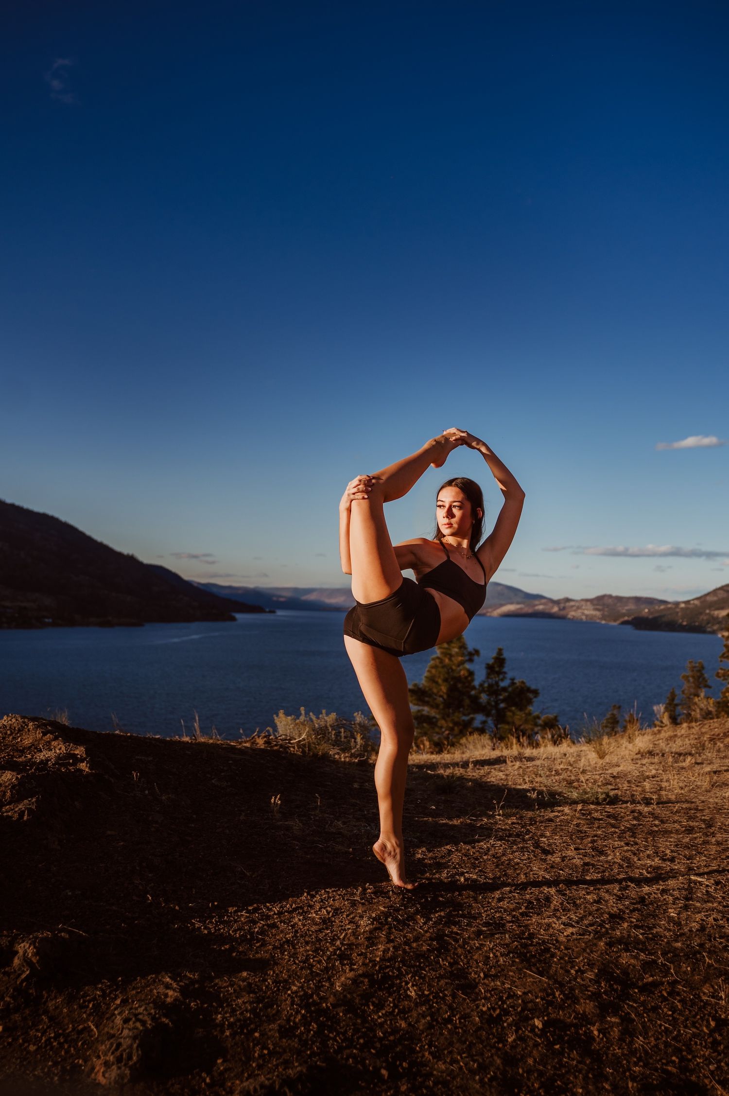 A dancer performs a standing split pose against a scenic mountain lake backdrop at sunset.