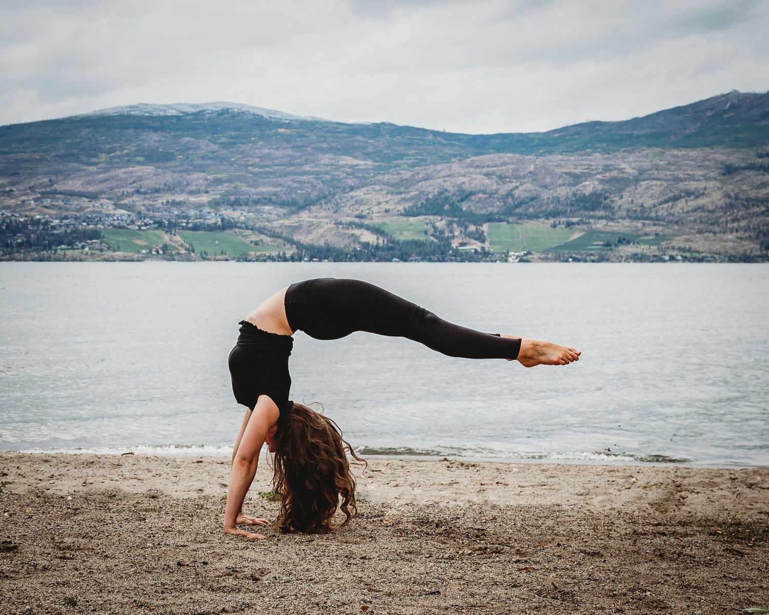 Gymnast performs graceful backbend on sandy beach with scenic mountain lake and hills in the background.