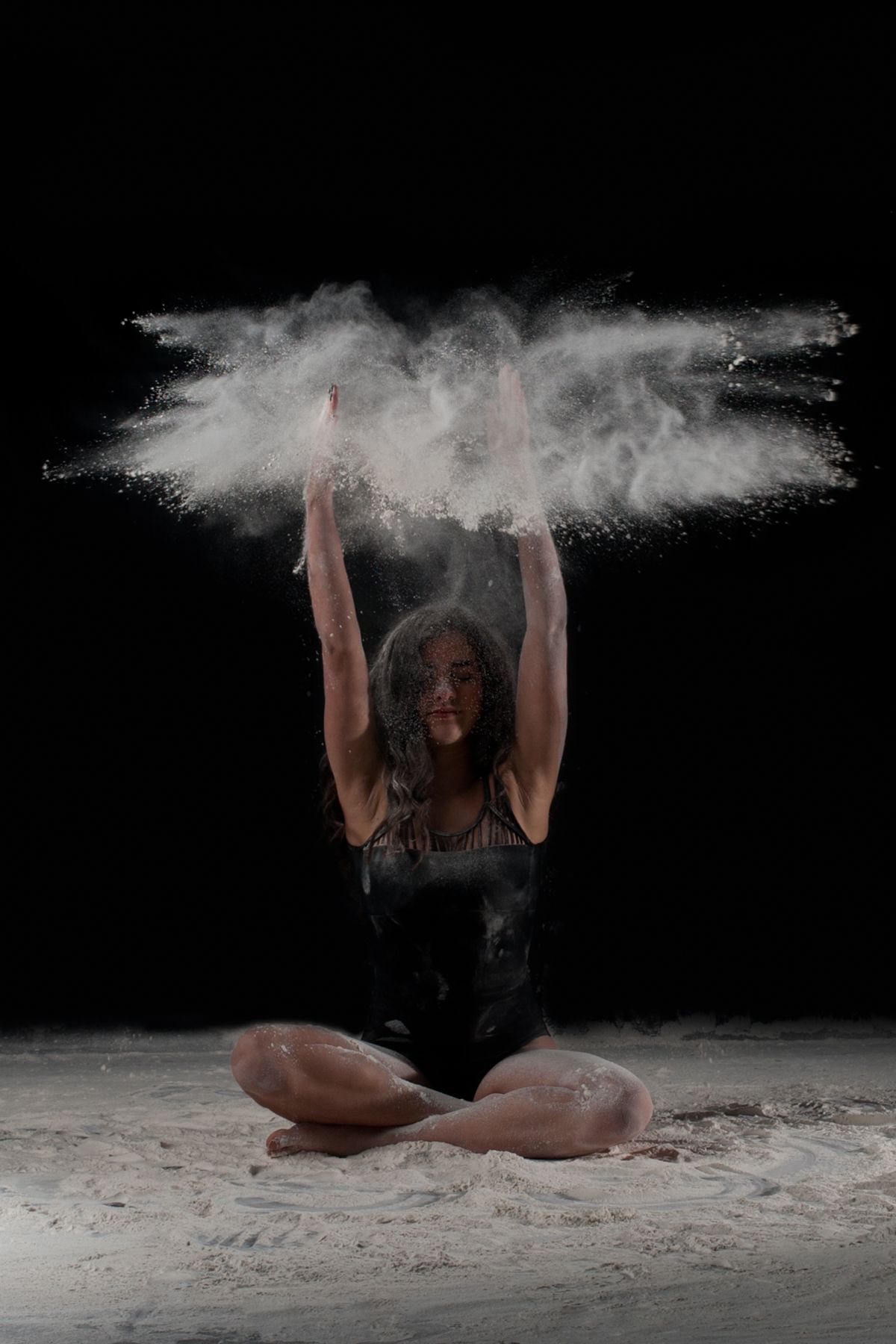 Contemporary dancer sits cross-legged creating dynamic white powder explosion effect above their head against black background.
