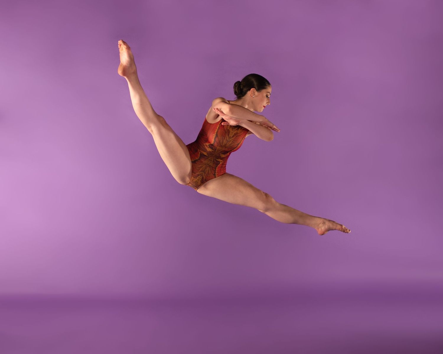 A gymnast in a red leotard performs a graceful split jump against a purple backdrop during a dance routine.