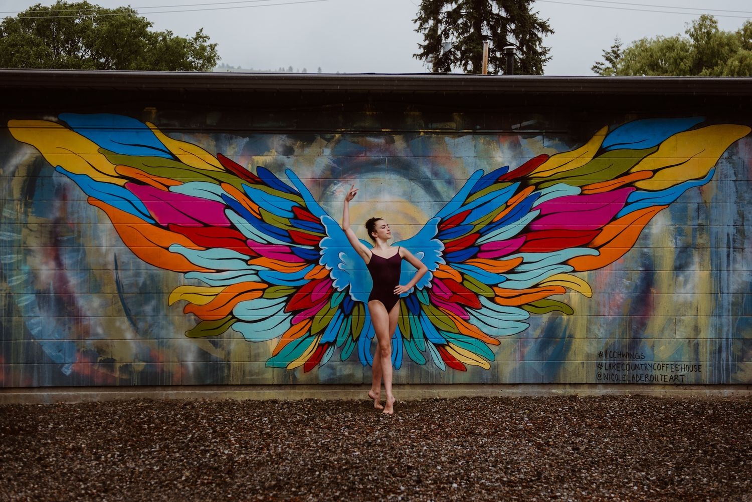 A dancer poses in front of vibrant street art mural featuring colorful geometric wing designs on a concrete wall.