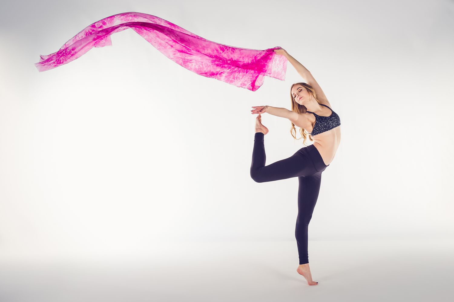 Dancer in black leggings poses gracefully with flowing pink silk fabric against white background.