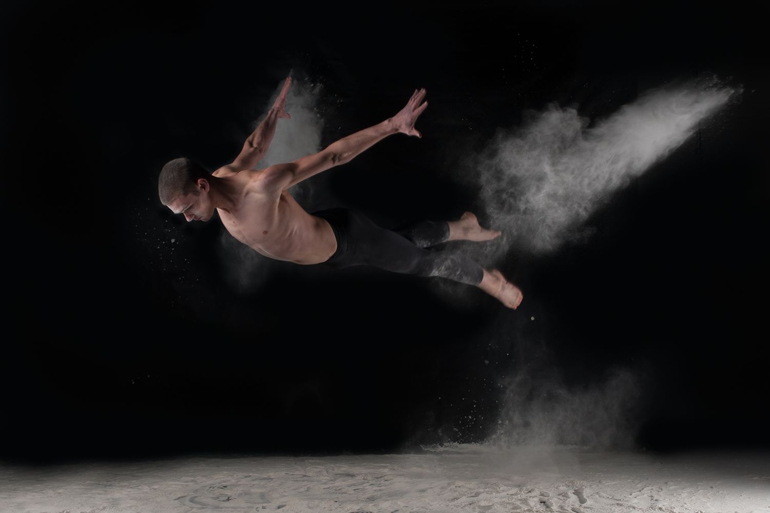 Dancer leaps gracefully through cloud of white powder against black background in dramatic artistic performance.