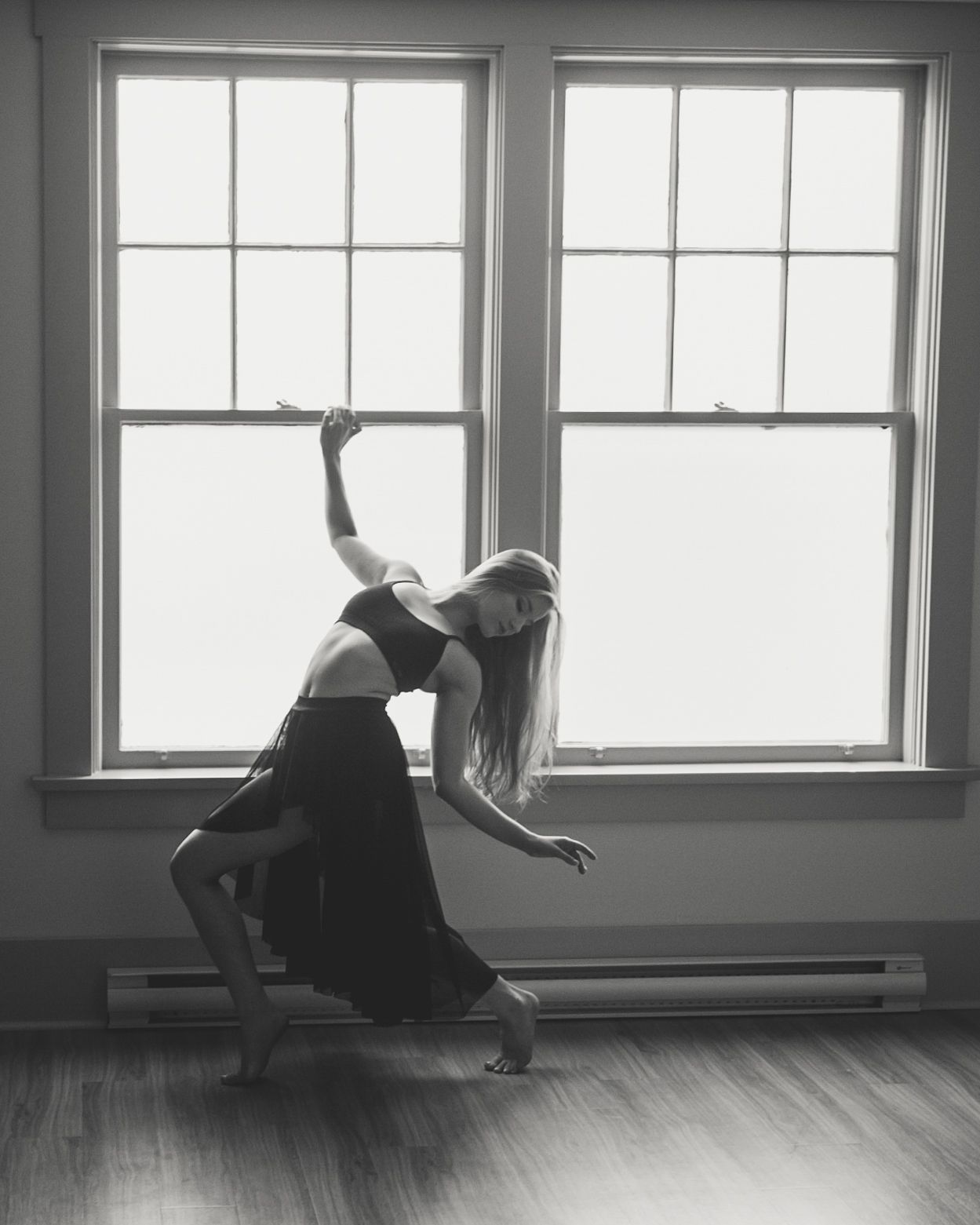 Dramatic black and white photo of a dancer in elegant pose near bright windows on hardwood floor.