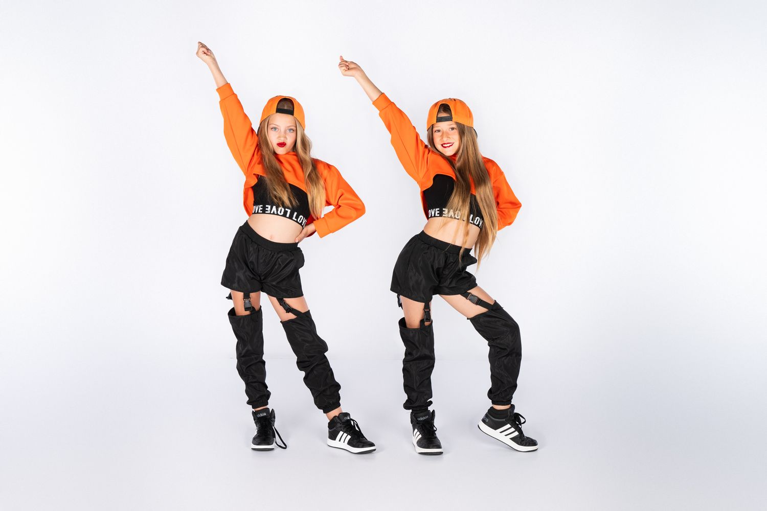 Two dancers in orange tops and black pants perform urban dance moves in studio setting.