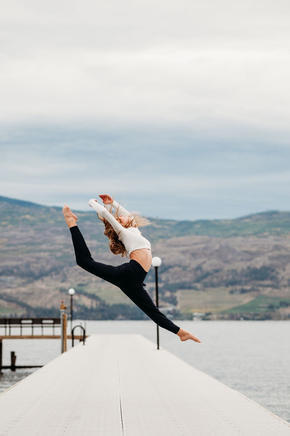 Dancer performs graceful leap over snowy dock with mountain lake and hills in background.