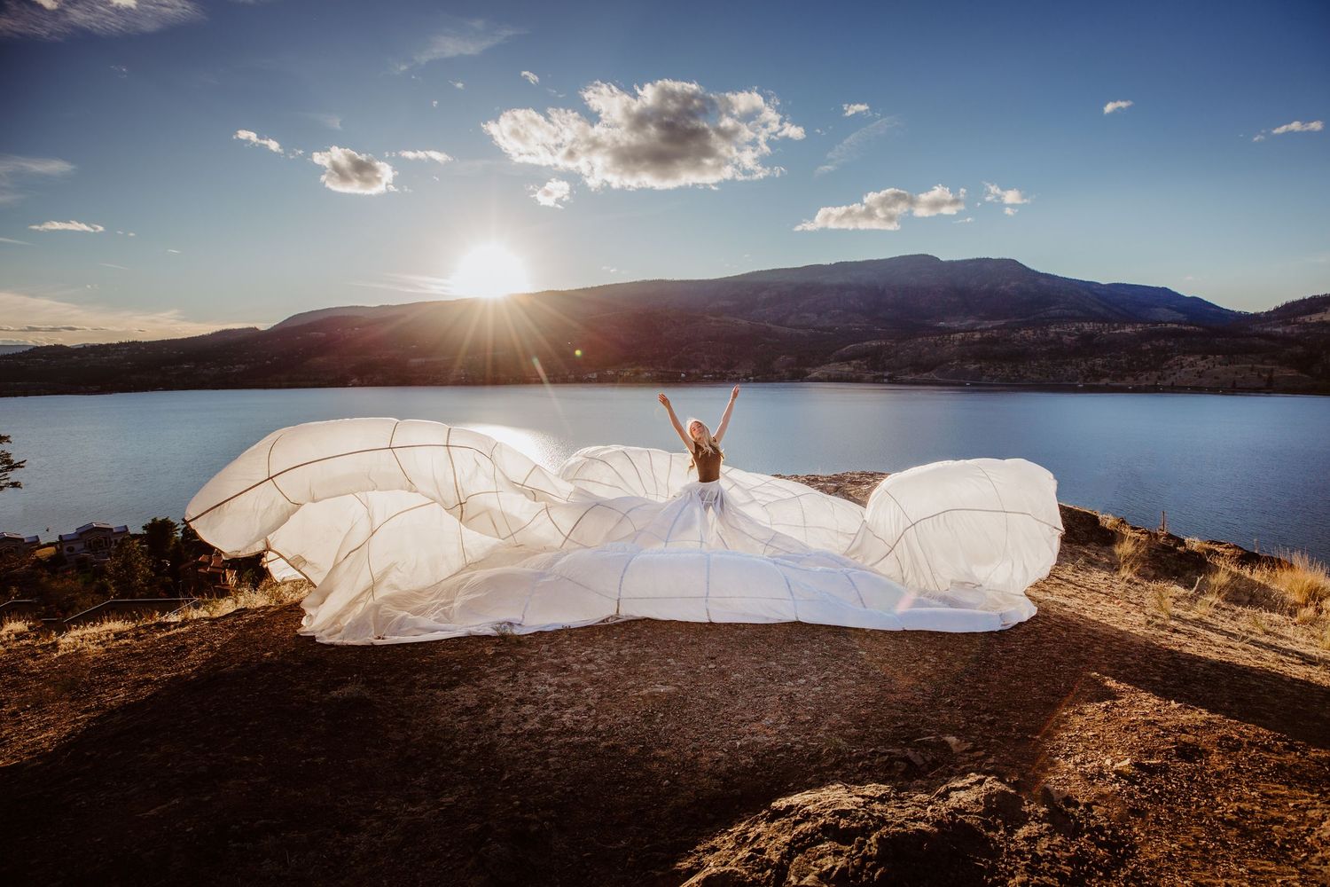 A flowing white wedding dress billows dramatically against a scenic mountain lake backdrop at sunset.