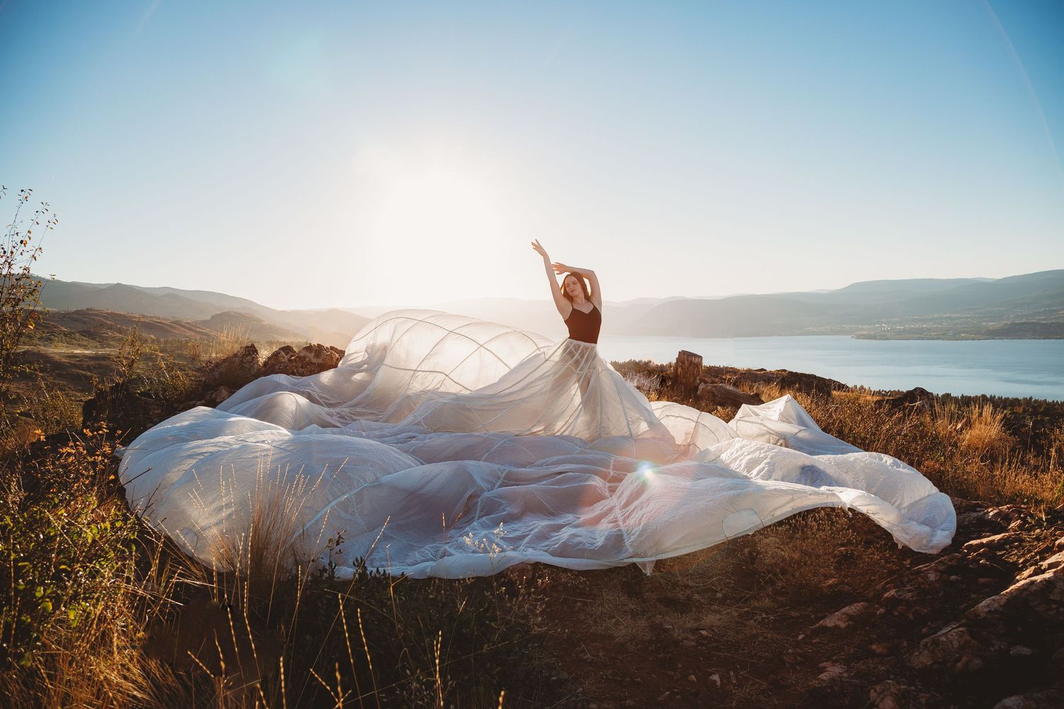 A flowing white wedding dress billows dramatically against a scenic mountain lake backdrop at sunset.