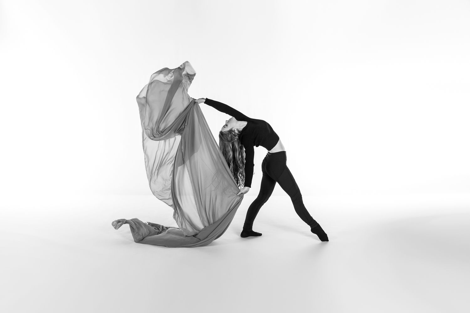 A dancer in black leotard performs a dynamic pose while holding flowing white fabric against a white background.