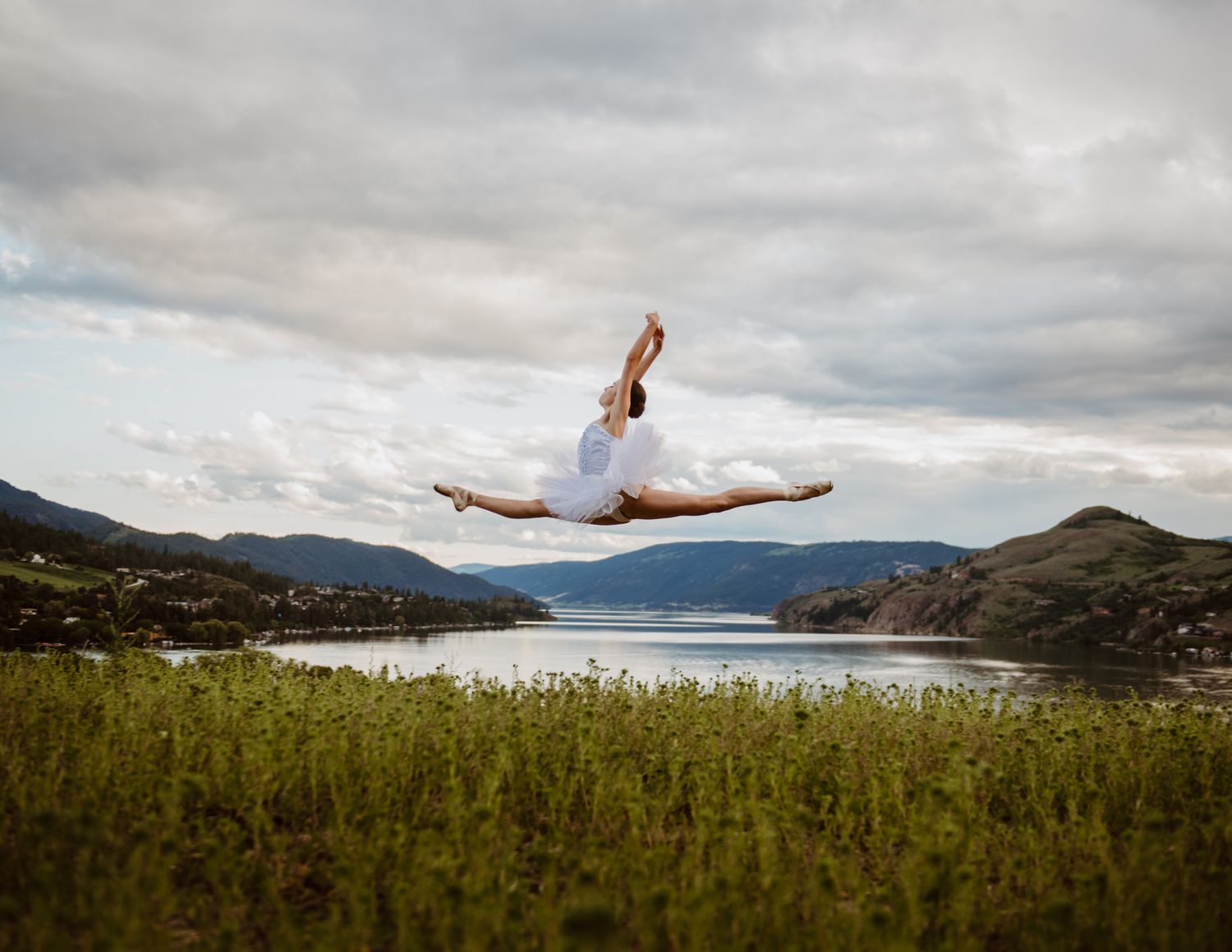 Dancer performs a dramatic split leap against a scenic mountain lake backdrop under cloudy skies.