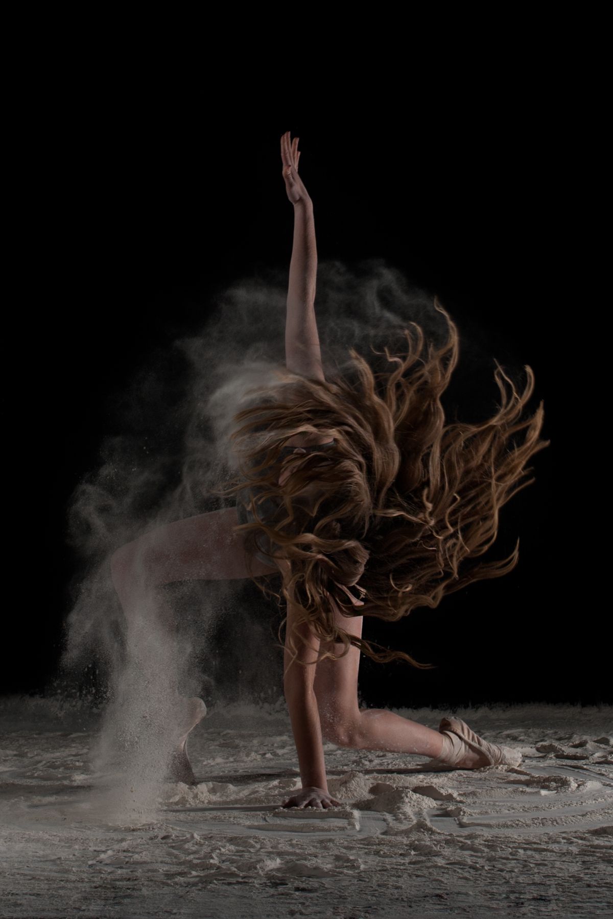Dramatic dance pose with flowing hair and flour dust creating ethereal effect against dark background.