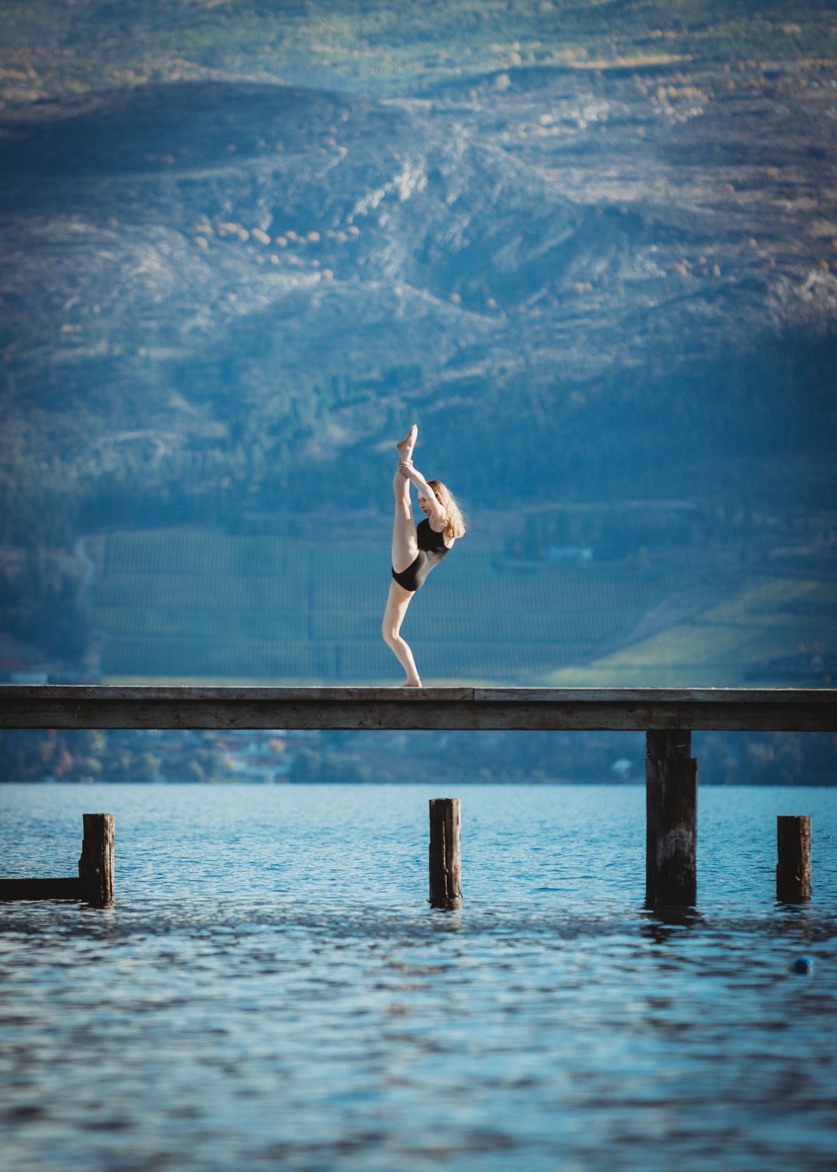 Graceful gymnast performs a ballet-inspired pose on a wooden dock overlooking a serene mountain lake.