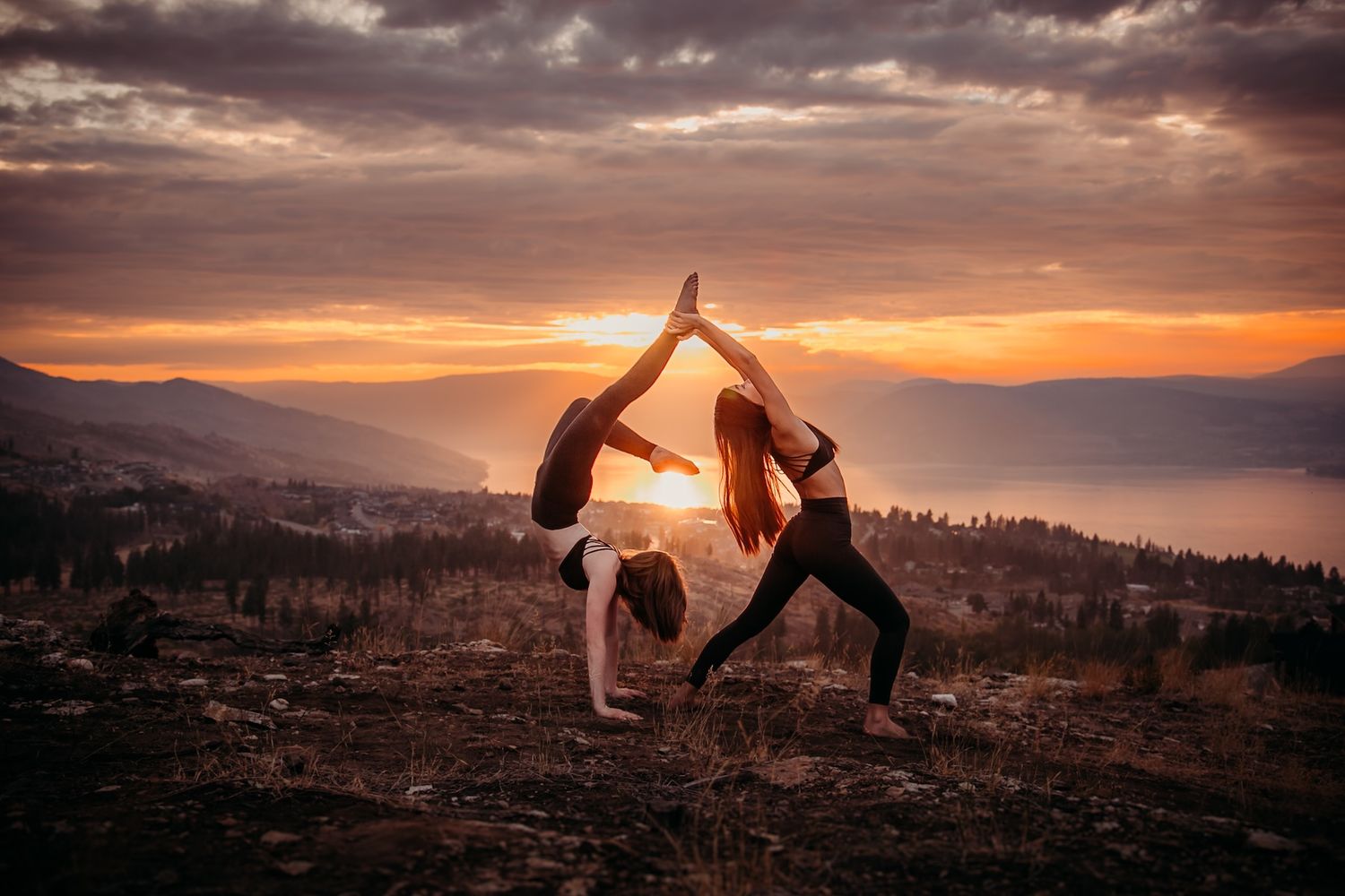 Two silhouetted figures perform an acrobatic yoga pose against a dramatic mountain sunset backdrop.
