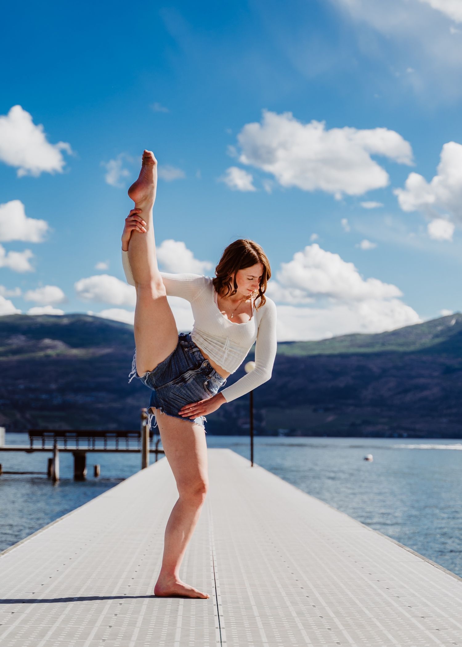 Athletic dancer performs a leg extension pose on a dock by a scenic mountain lake under blue skies with white clouds.