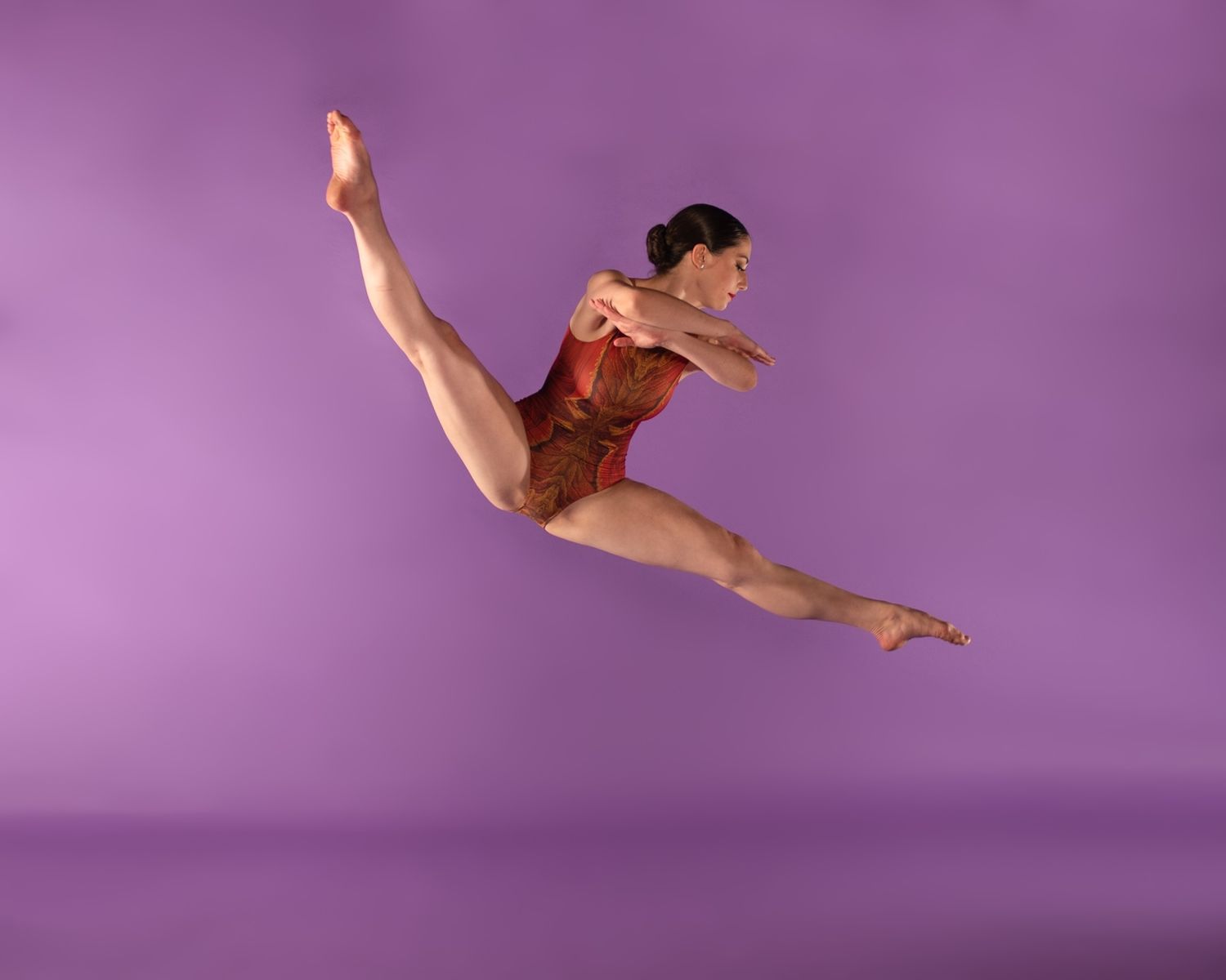 A gymnast in a red leotard performs a graceful aerial split leap against a purple backdrop during a dance performance.