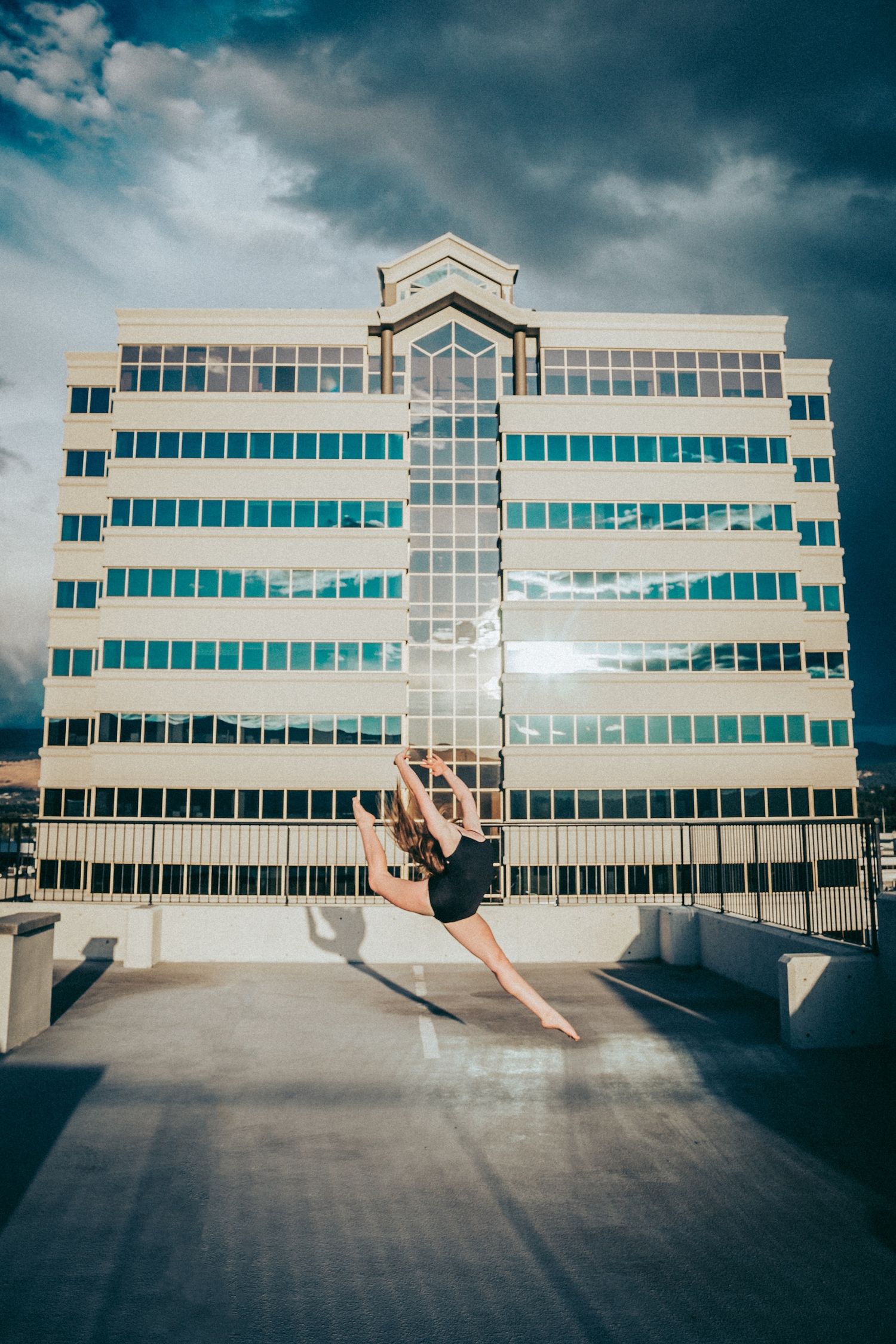 Dancer leaps gracefully on a rooftop against dramatic cloudy sky and modern office building backdrop.