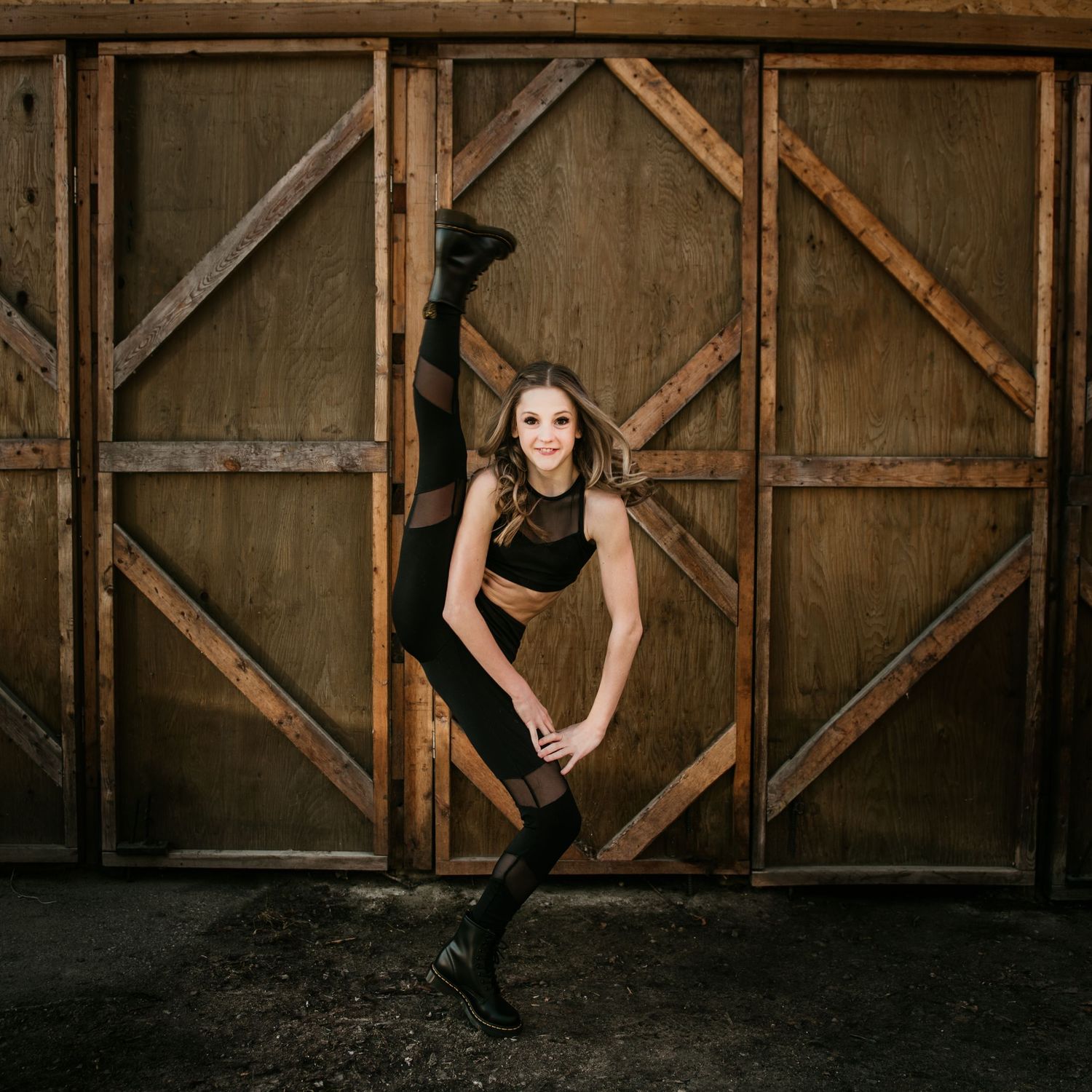 Dancer in black performs a dramatic leg split against rustic wooden barn doors in artistic photoshoot.