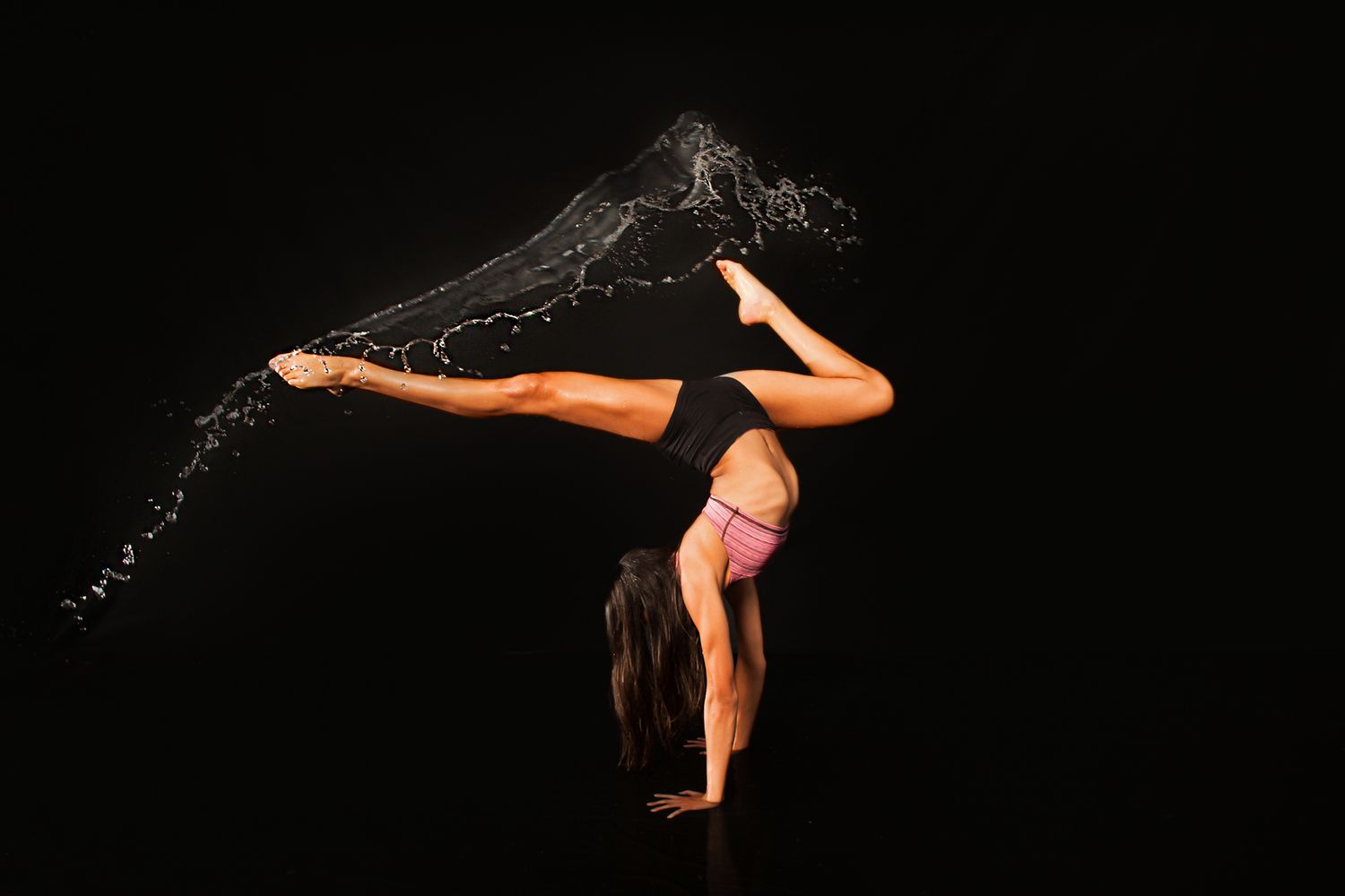Gymnast performing handstand with water splash effect creating artistic patterns on black background.