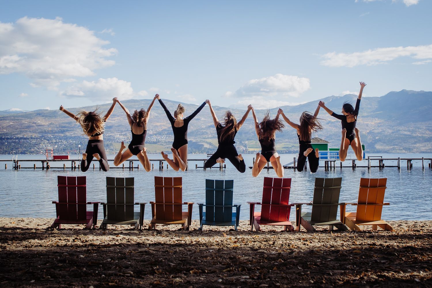 Six friends jump joyfully from colorful Adirondack chairs on a beach with mountains and lake in the background.