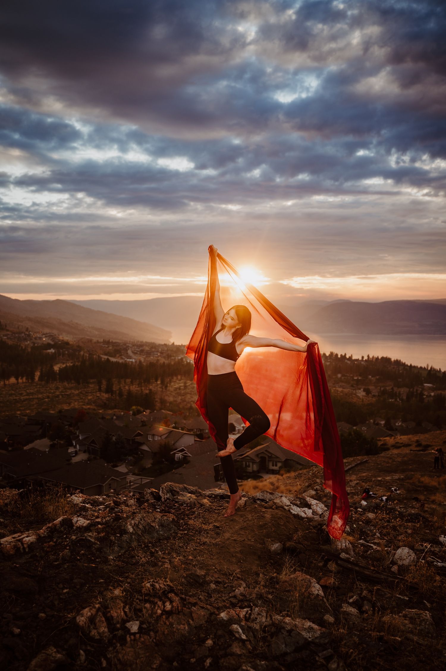 Dancer poses with flowing red fabric against dramatic mountain sunset landscape.