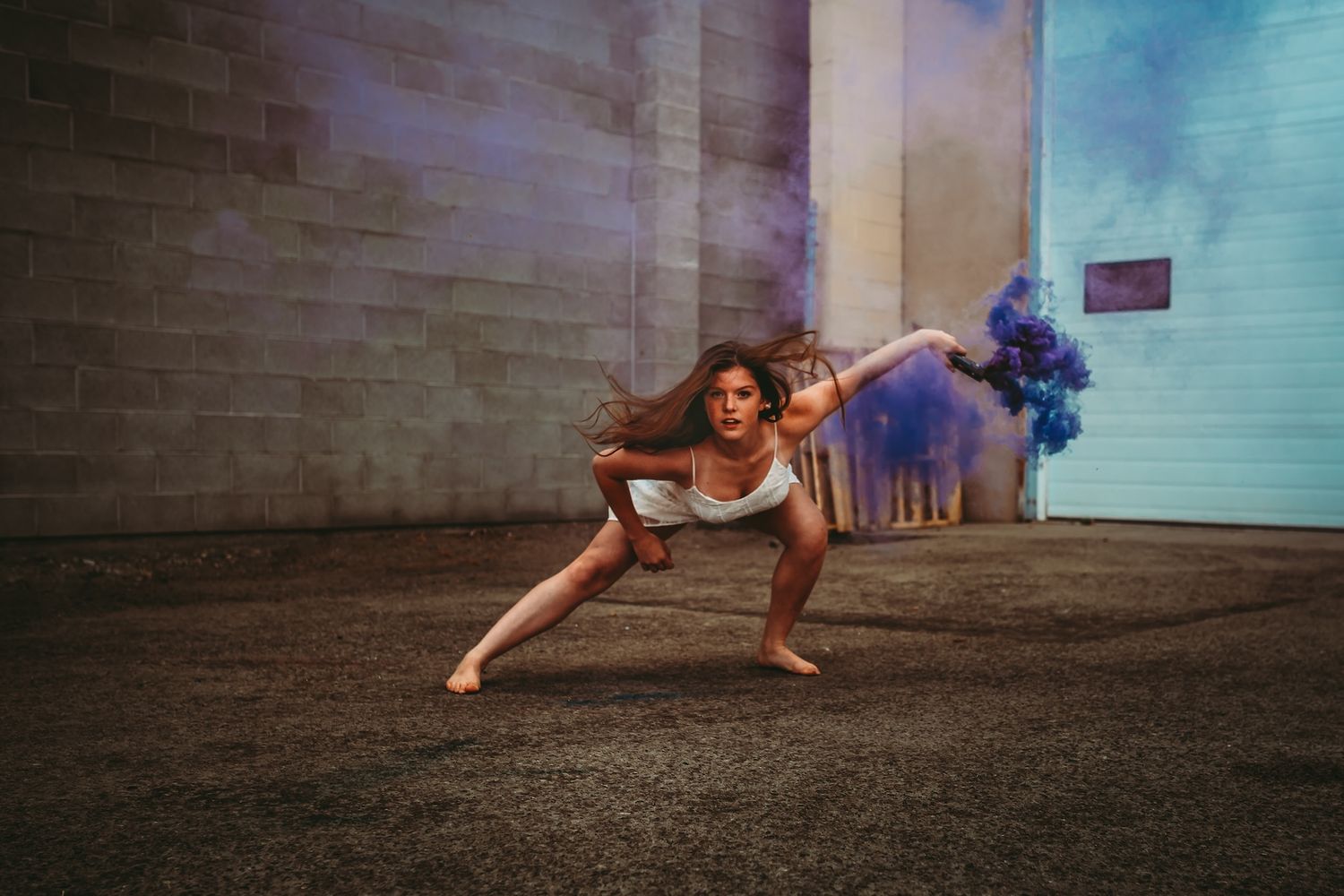 Dancer in beige dress performs dramatic pose with blue smoke effects against urban wall backdrop.