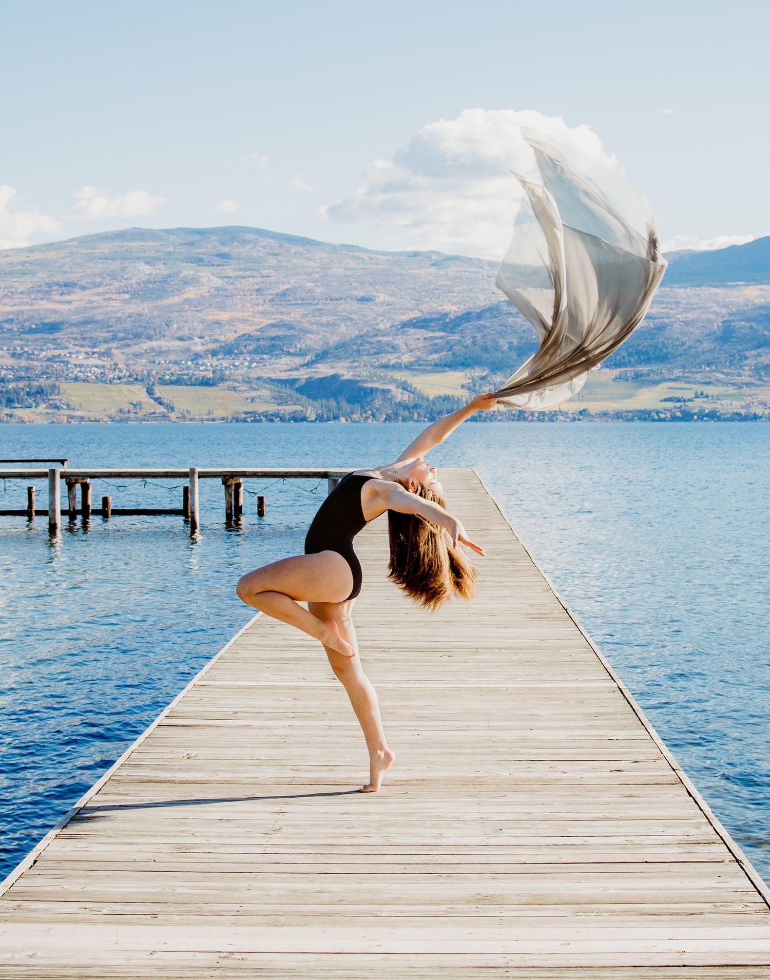 Dancer performs graceful pose with flowing fabric on wooden pier overlooking mountain lake on sunny day.