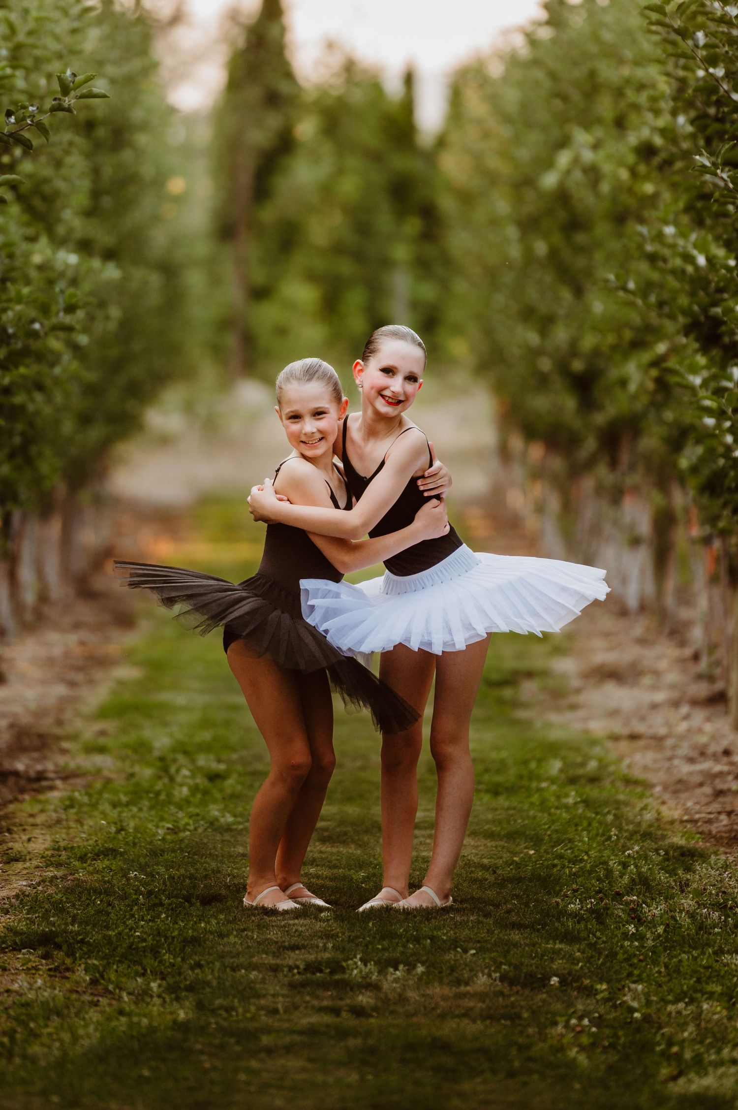 Two ballet dancers in leotards and tutus pose playfully together between rows of trees in an orchard.