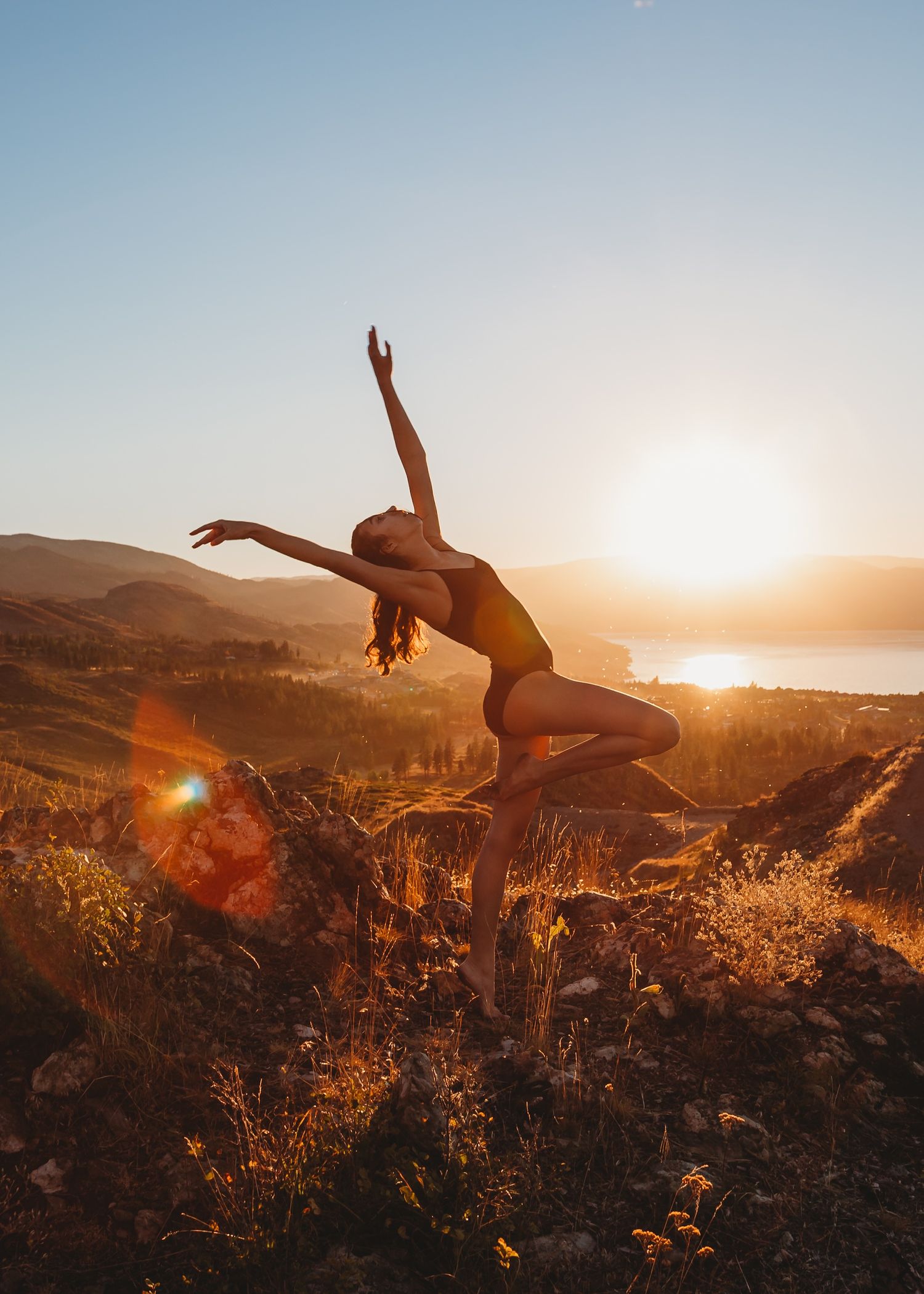 Dancer performs graceful leap against golden sunset over coastal hills and ocean landscape.