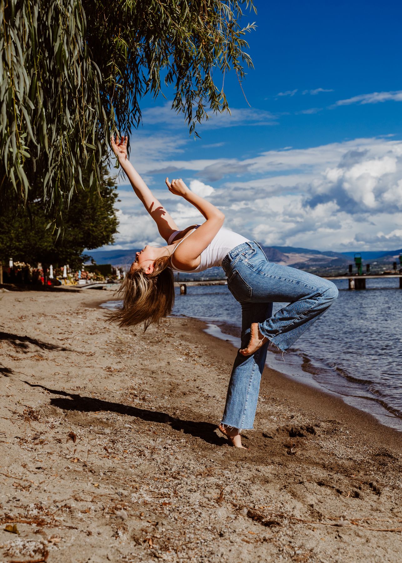 A dancer performs an elegant backbend pose on a sunny beach with mountain and lake views in the background.