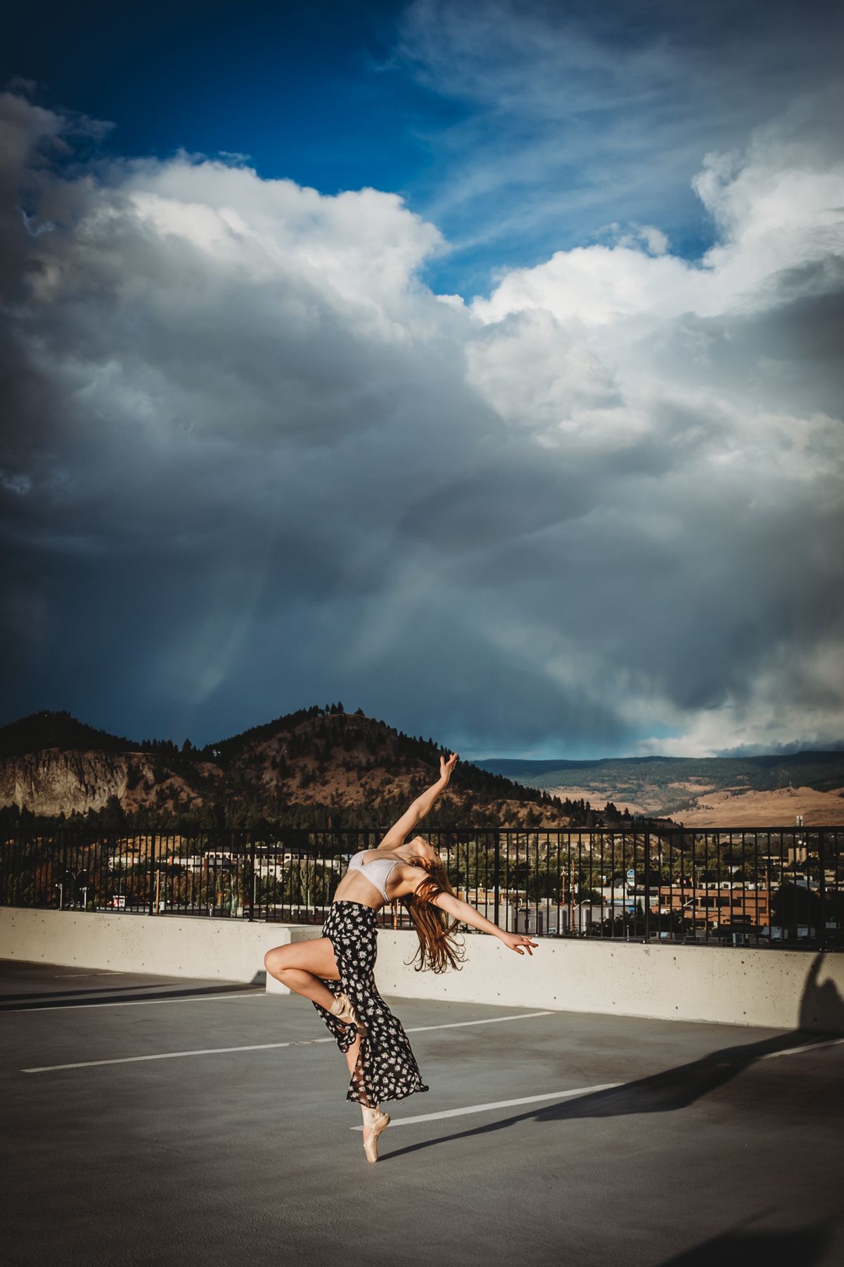 A dancer leaps gracefully in a flowy polka dot dress against a dramatic mountain landscape with stormy clouds overhead.