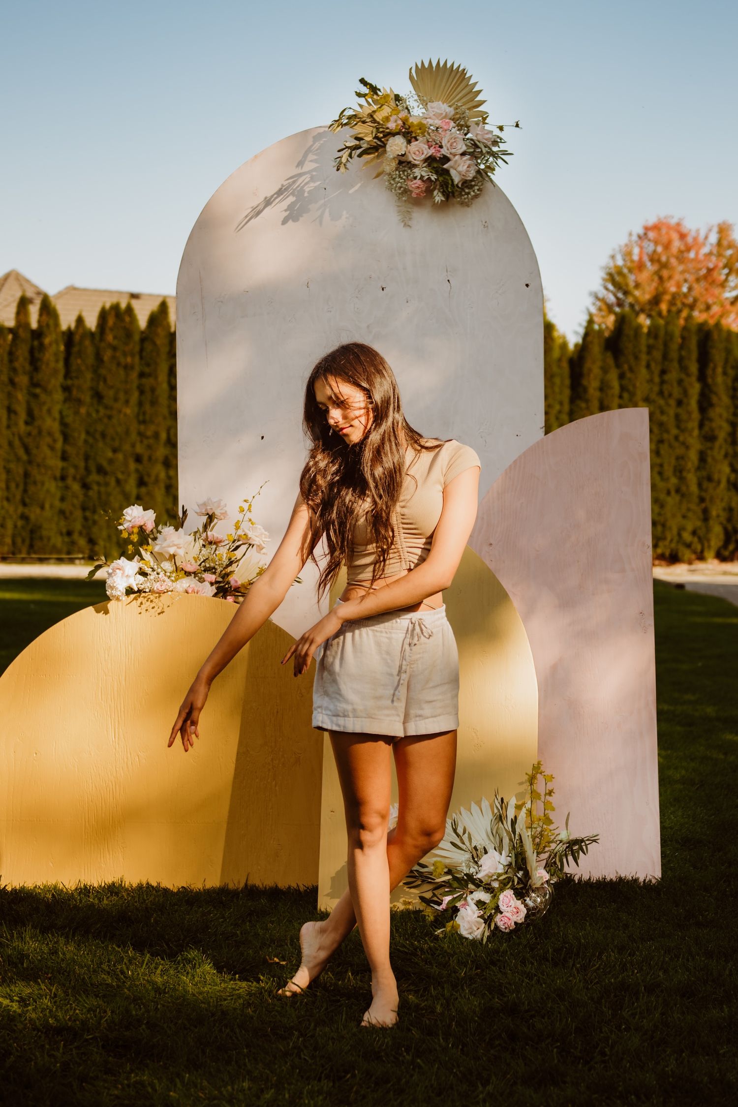 A figure in casual summer attire poses against a curved modern backdrop with floral arrangements at sunset.