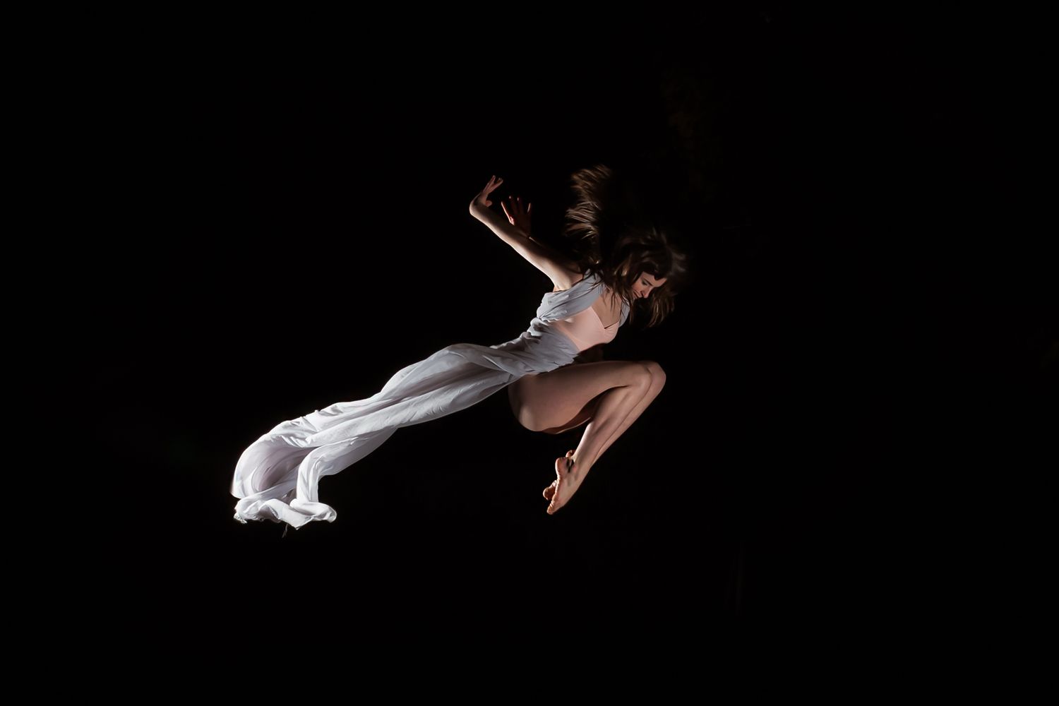 Dramatic dancer leaps through darkness wearing flowing white fabric against black background.