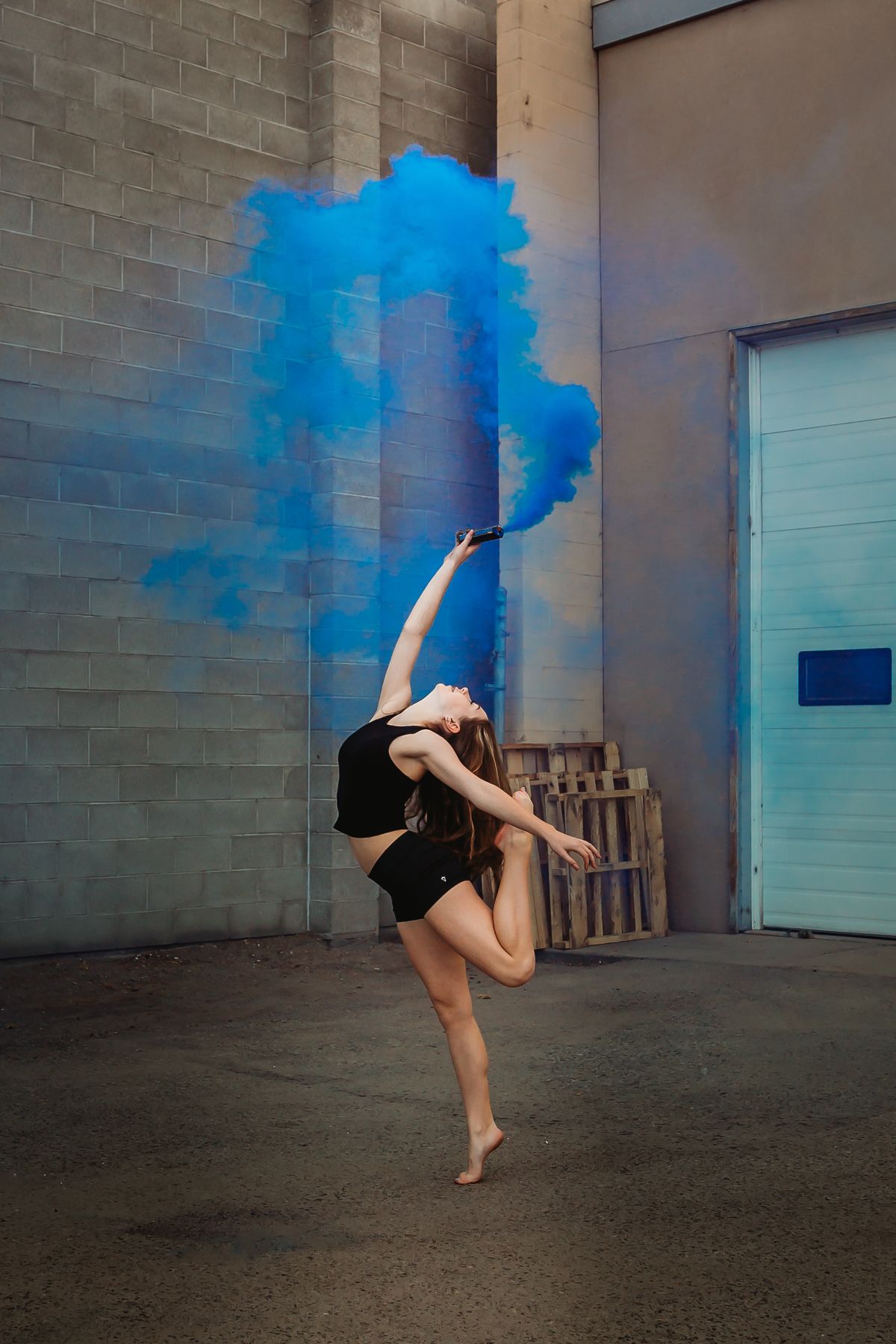 Dancer in black performs graceful leap pose against industrial wall with vibrant blue smoke effect.