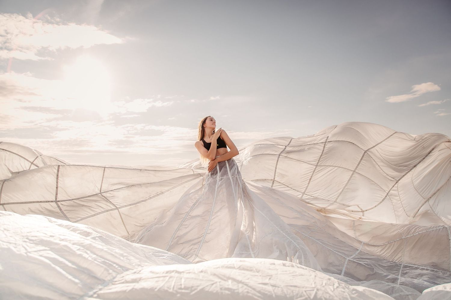 A person in flowing white fabric stands against a bright sky as ethereal fabric billows dramatically in the wind.