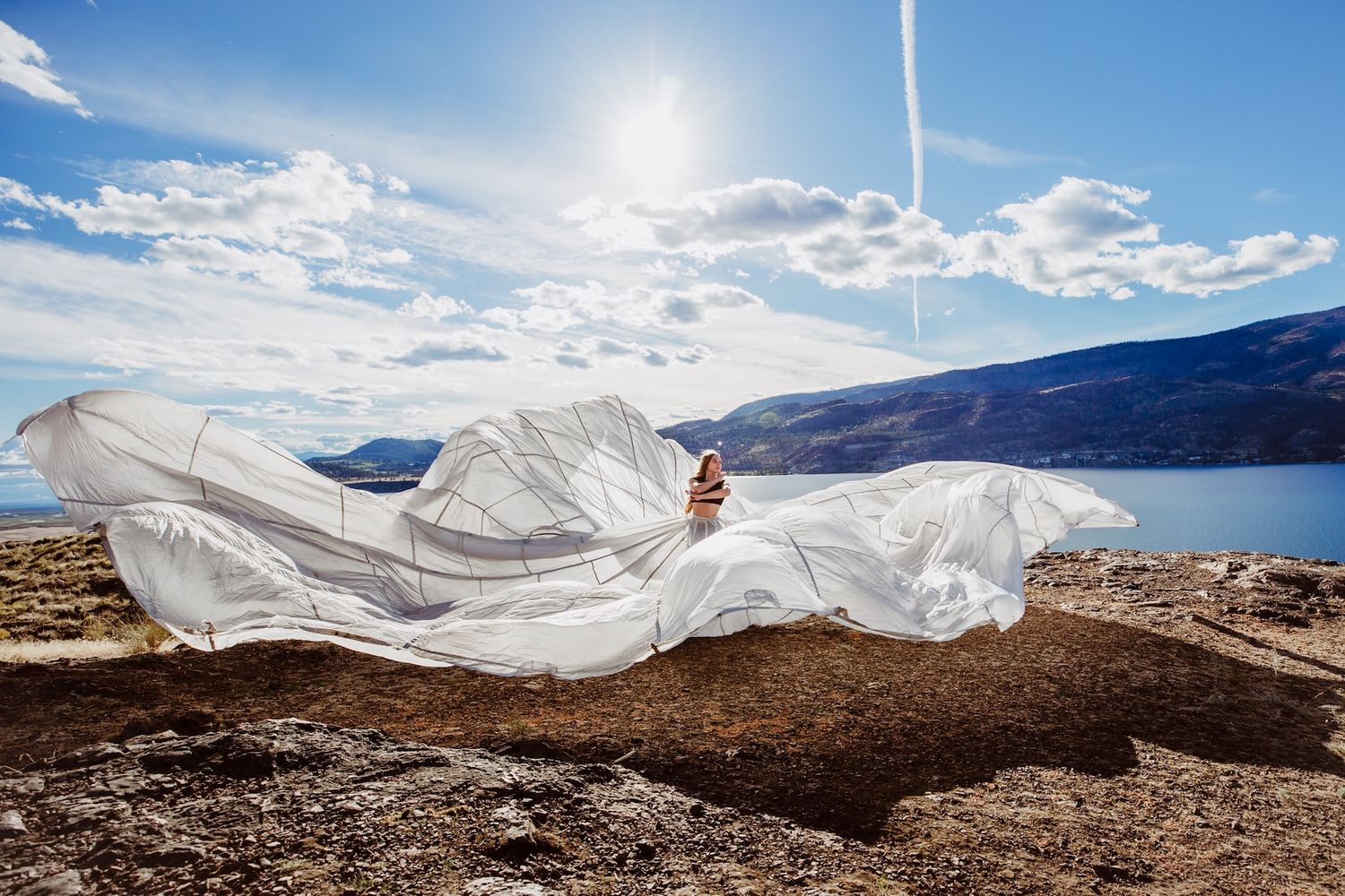 White flowing fabric billows dramatically against a scenic mountain landscape with blue lake and bright sunlit sky.