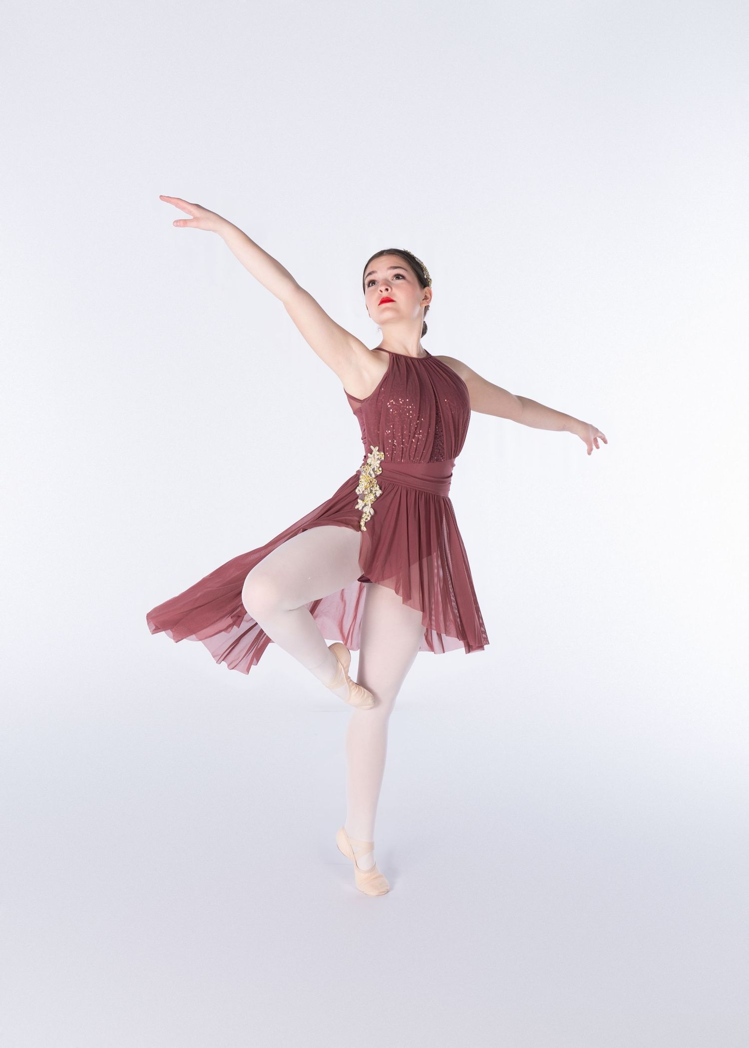 Ballet dancer in burgundy dress performs graceful pose on pointe in elegant studio setting against white background.