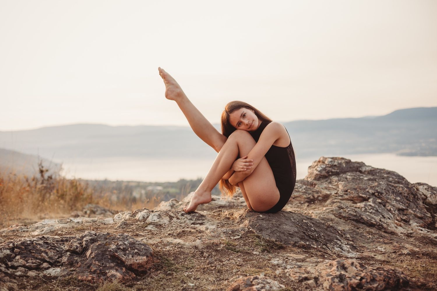 Athletic woman in black workout attire sits and points while posing on rocky cliff overlooking scenic mountain landscape.