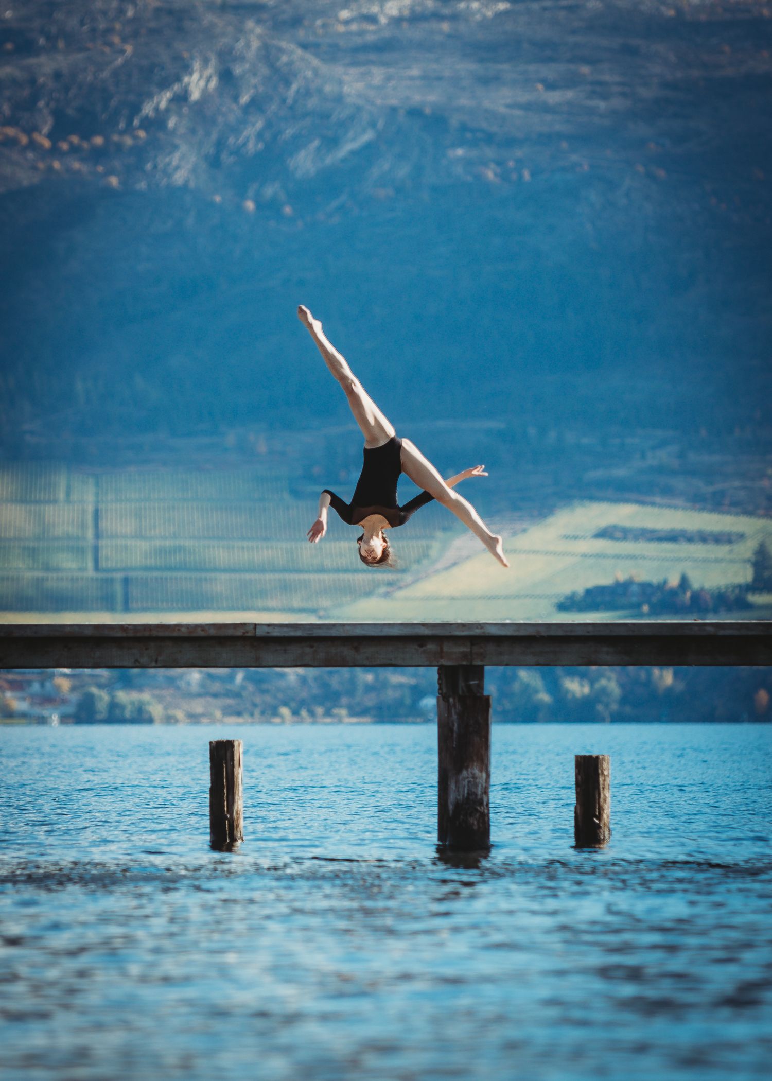 A person performs an athletic backflip off a wooden pier over a serene lake surrounded by mountains.