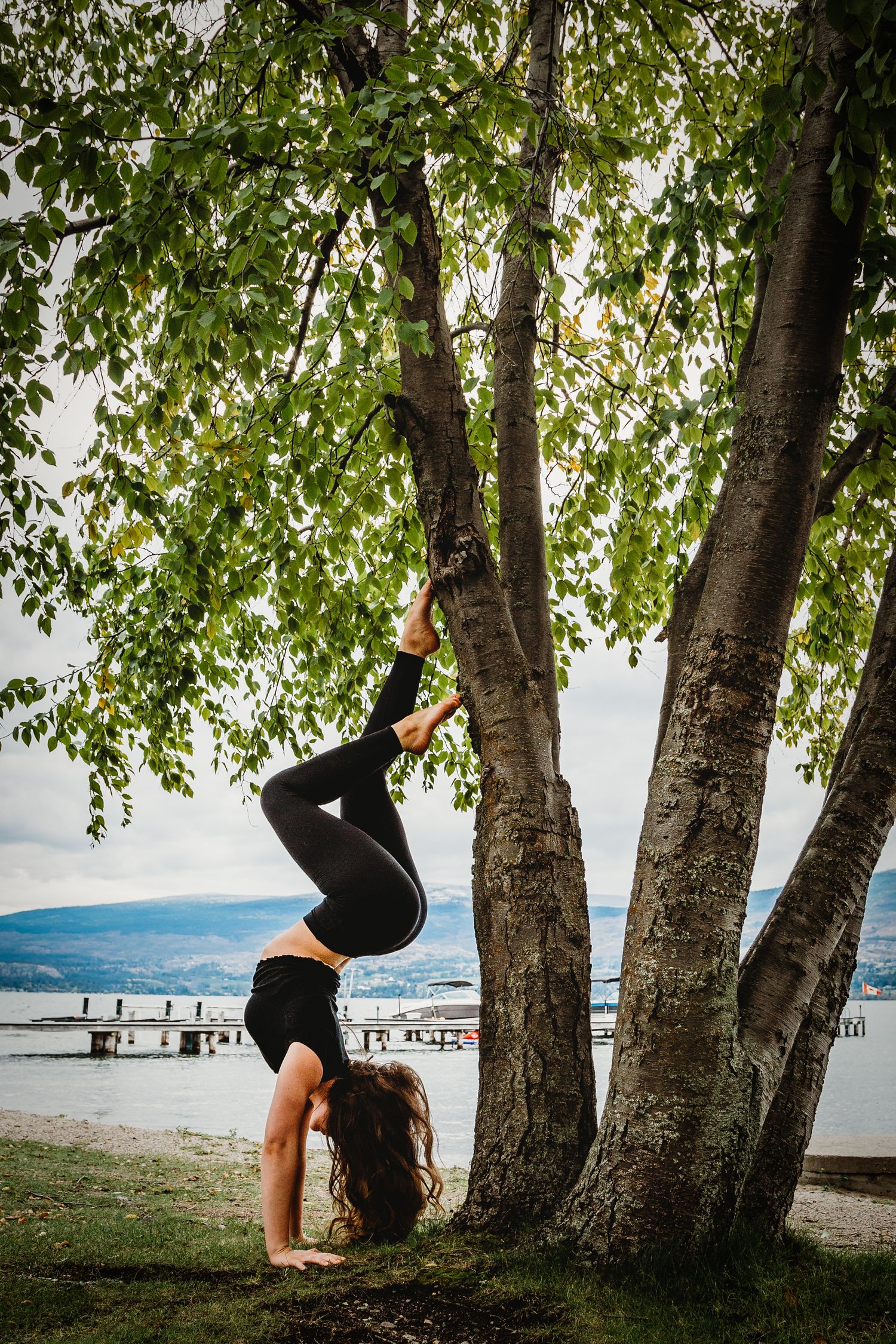 A gymnast performs a handstand against a birch tree by a lake with mountains in the background.